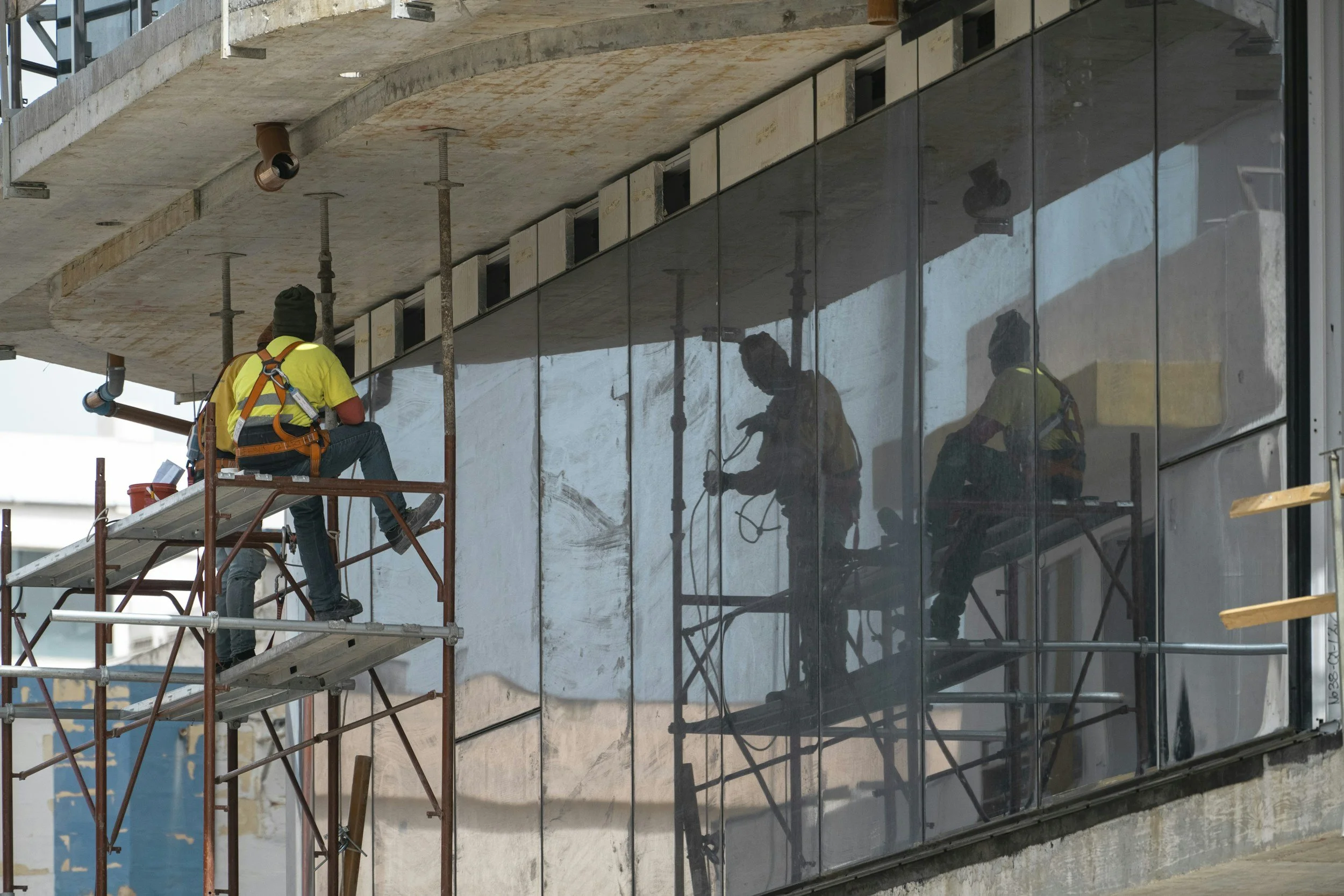 Construction workers installing large glass panels on the exterior of a building. The workers are wearing safety harnesses and yellow shirts, standing on scaffolding, with some working directly on the glass surface.