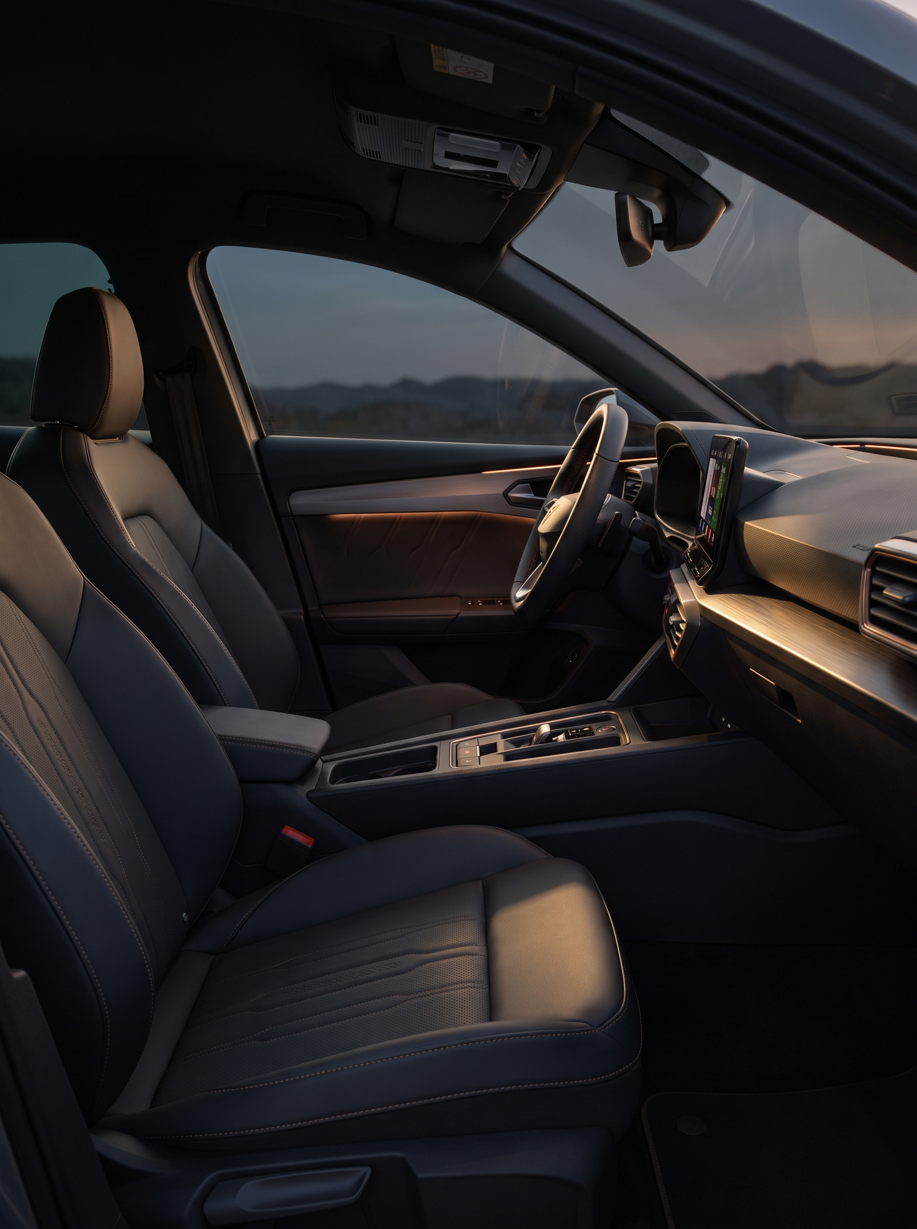 The interior of a modern car, showing the front passenger and driver seats, dashboard, and steering wheel with a large touchscreen display, in front of a landscape with hills at dusk.