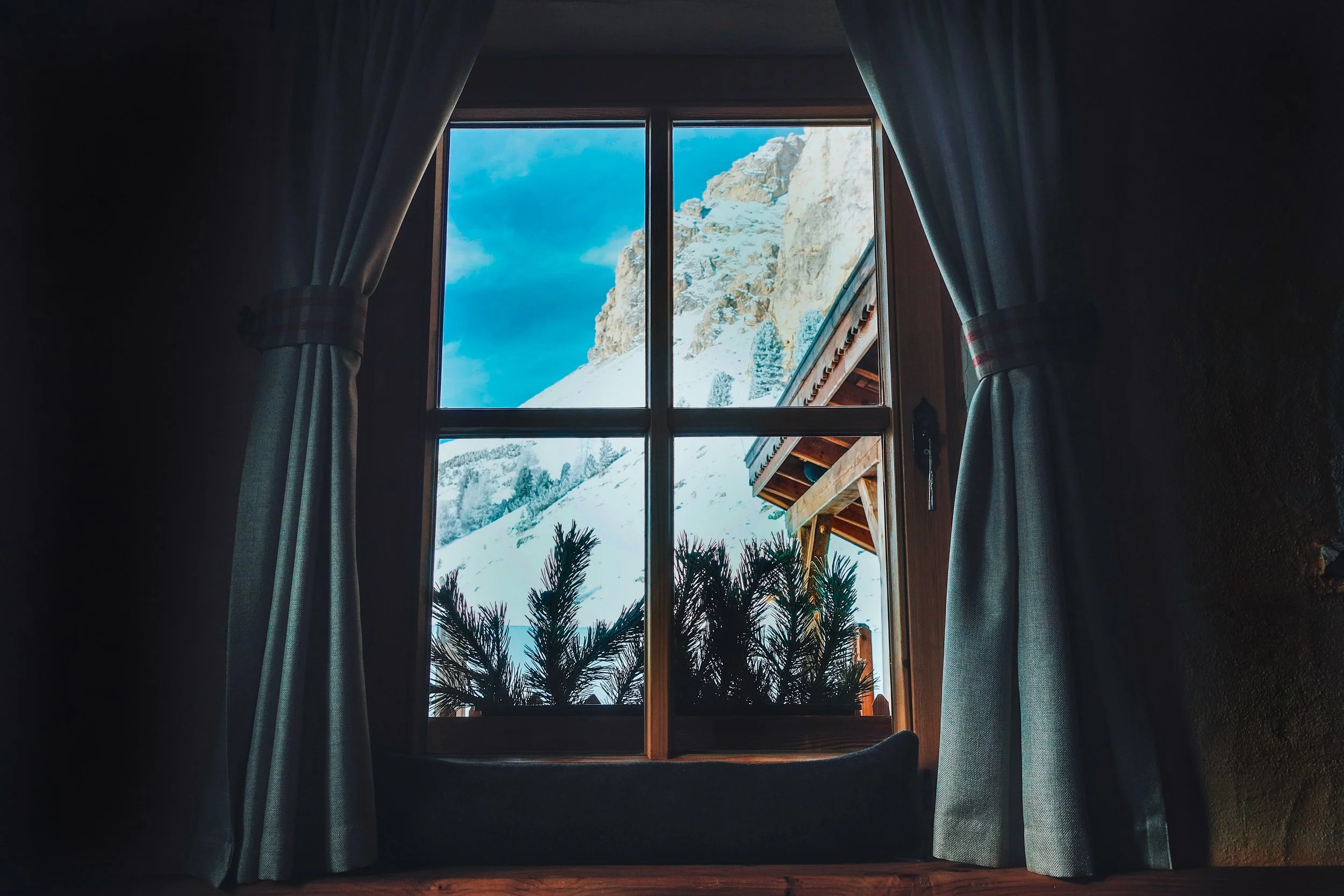 View through a window showing snow-covered mountains, blue sky, and clouds, framed by dark curtains inside a room.