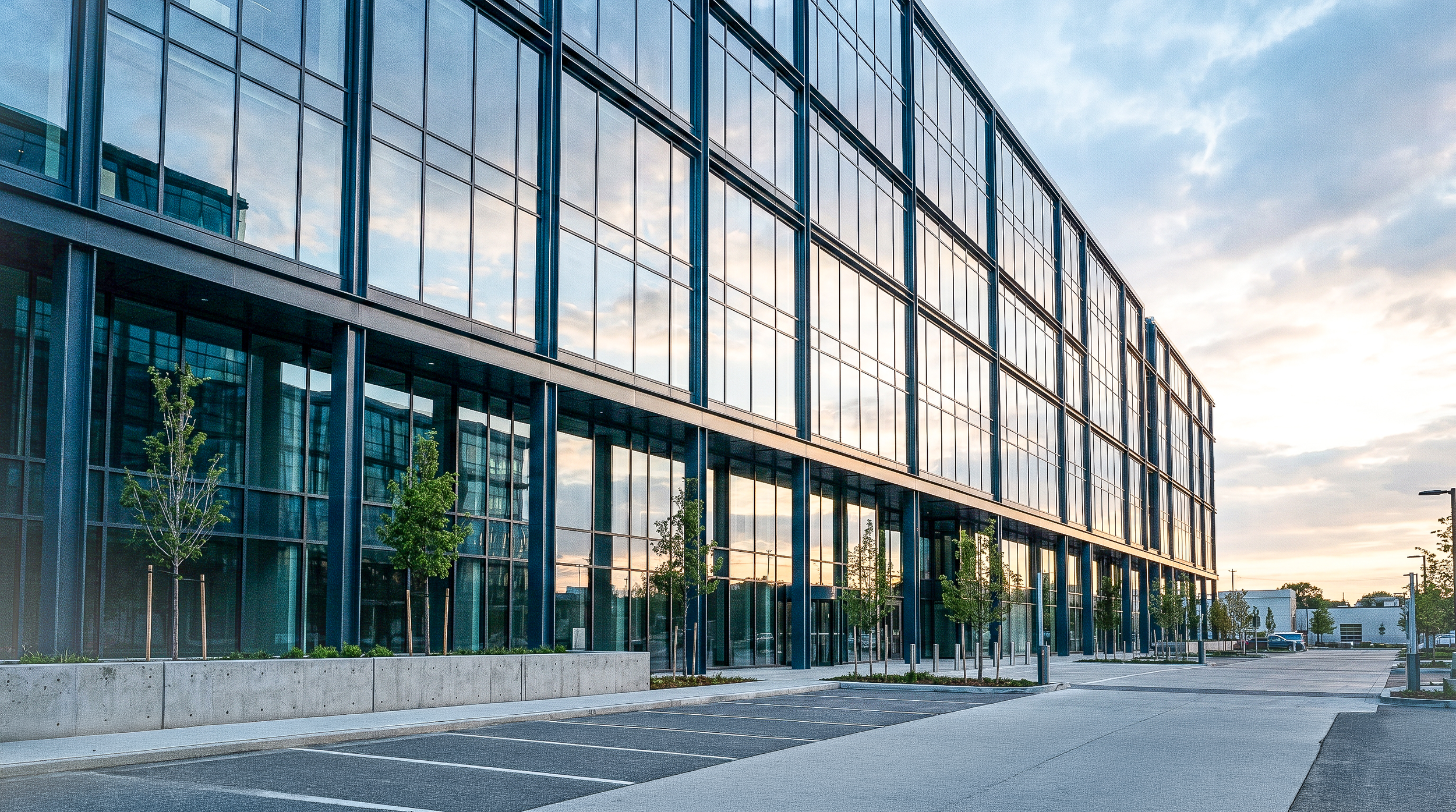 Modern glass building with reflections of the sky and trees, situated in an urban area with a parking lot, new trees, and streetlights.