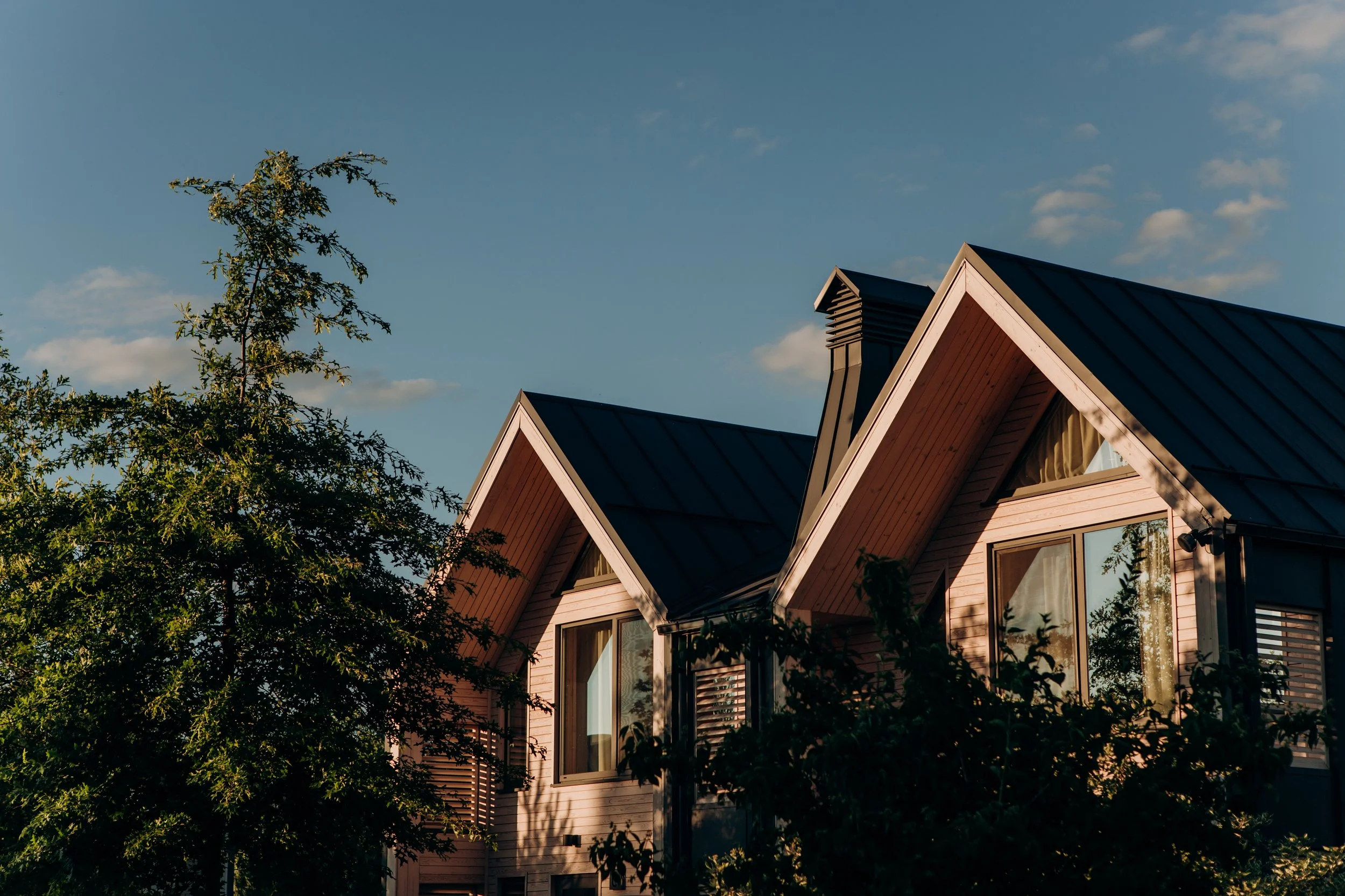 A modern, wooden house with a dark metal roof, surrounded by green trees, under a bright blue sky with scattered clouds.