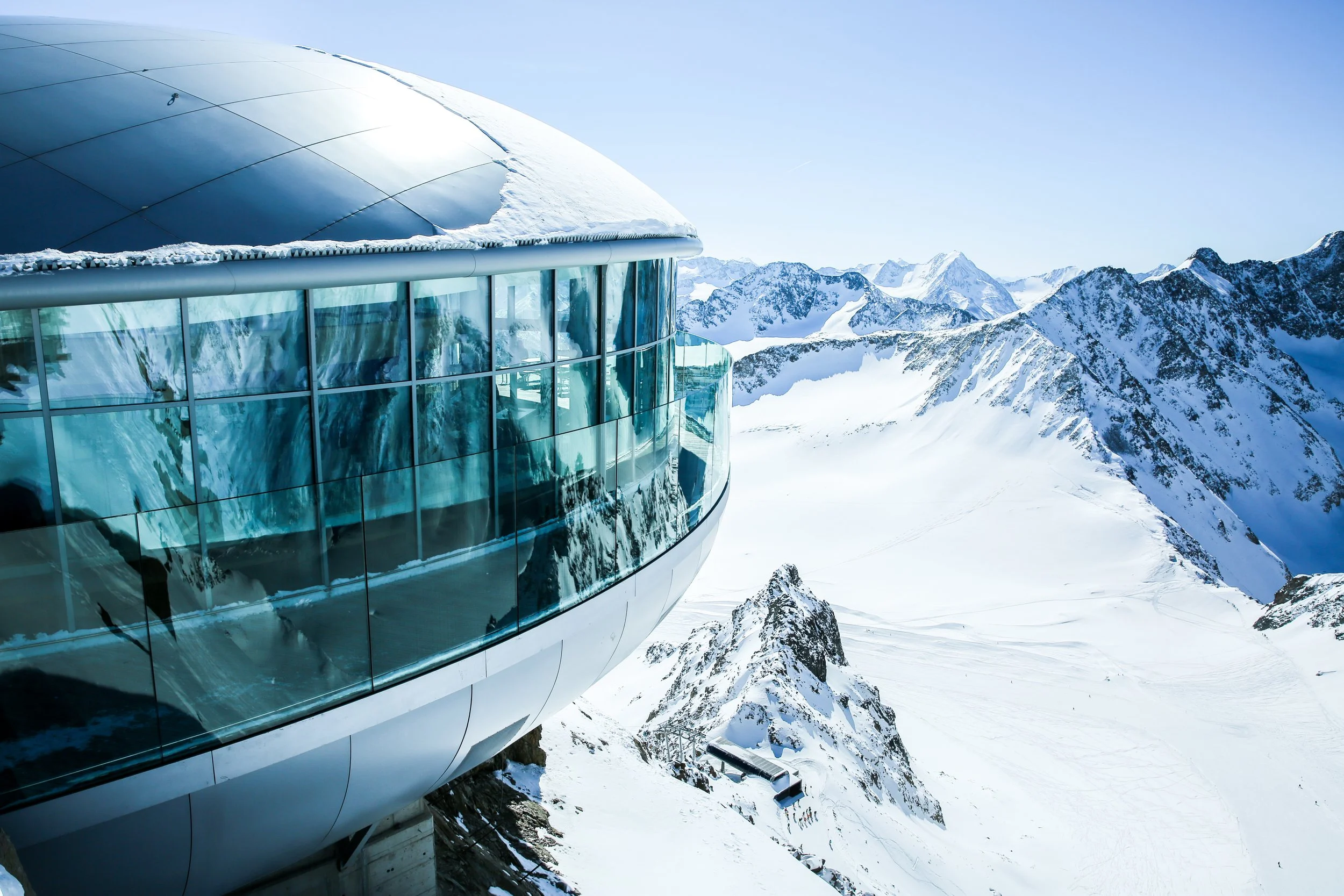 A modern glass building situated on snow-covered mountains with clear blue sky.