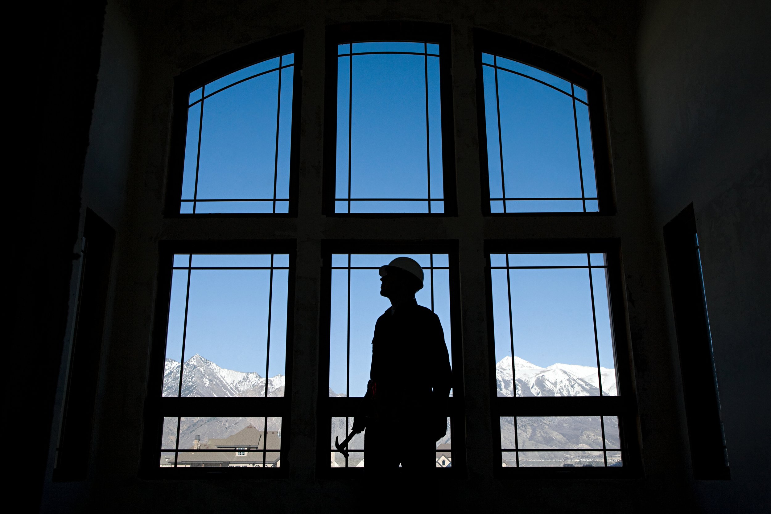 Silhouette of a construction worker with a helmet standing inside a building under construction, looking out the large window at snow-capped mountains in the distance.