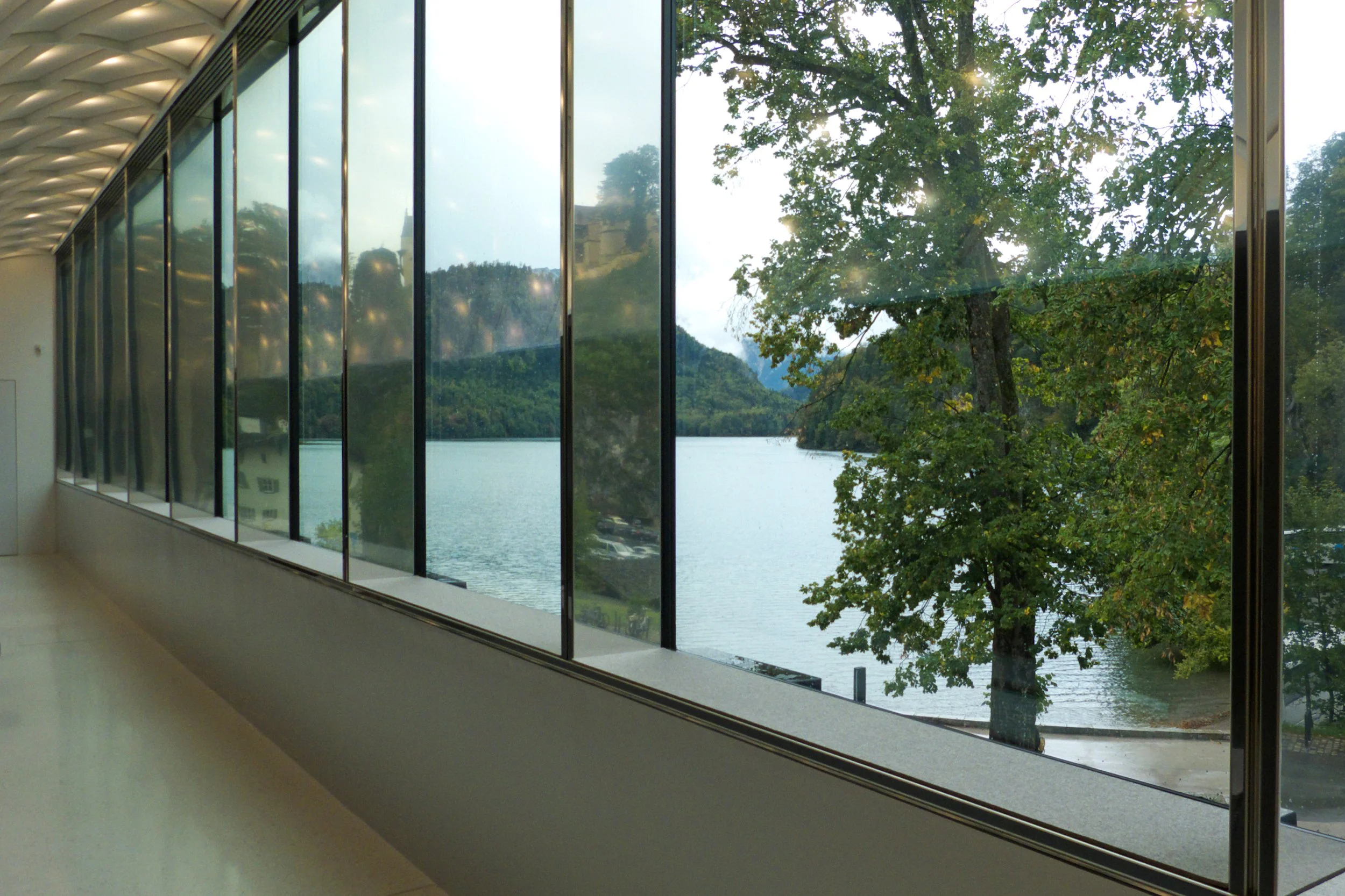 Interior view of a building with large floor-to-ceiling windows showing trees, a river, and mountains outside.
