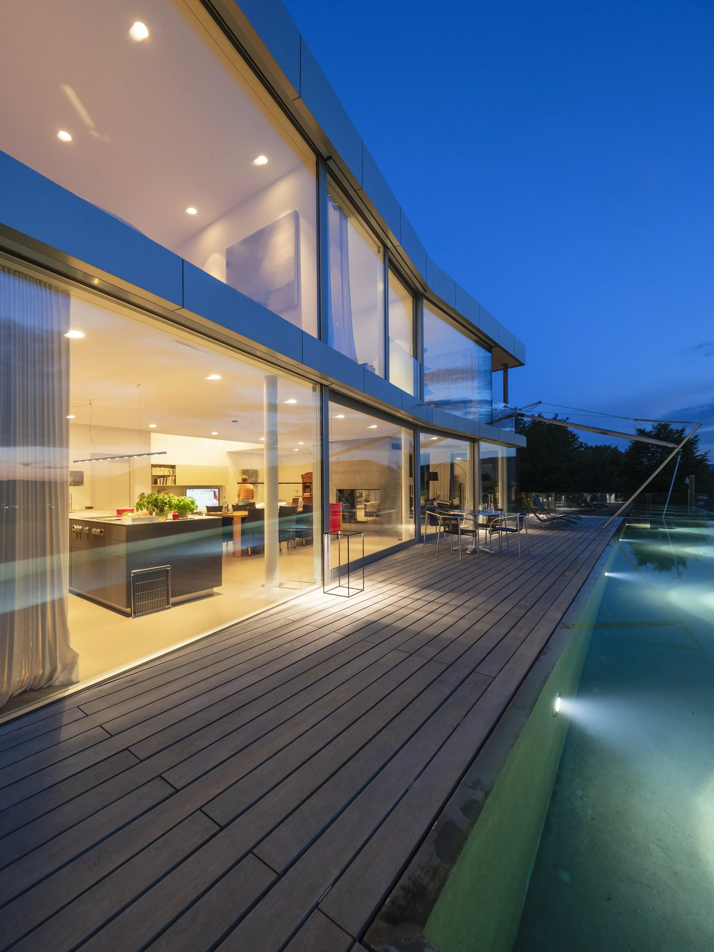 Modern house with large glass windows illuminated at night, wooden deck, outdoor seating, swimming pool, and a view of the blue twilight sky.