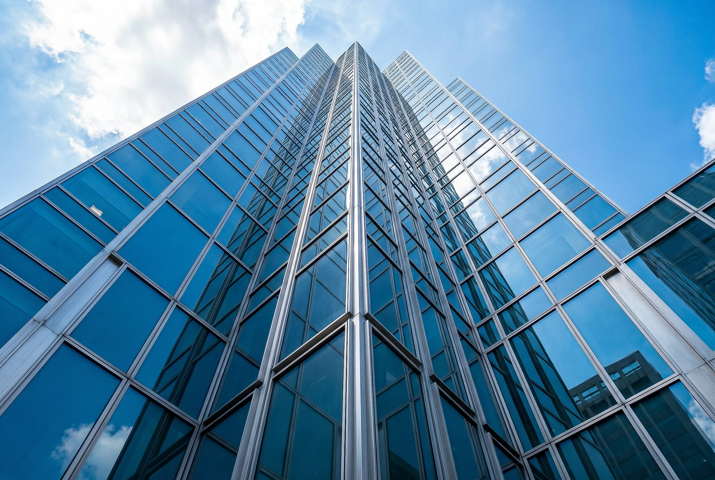 A modern glass skyscraper viewed from the ground looking up, with a blue sky and clouds reflected on its surface.