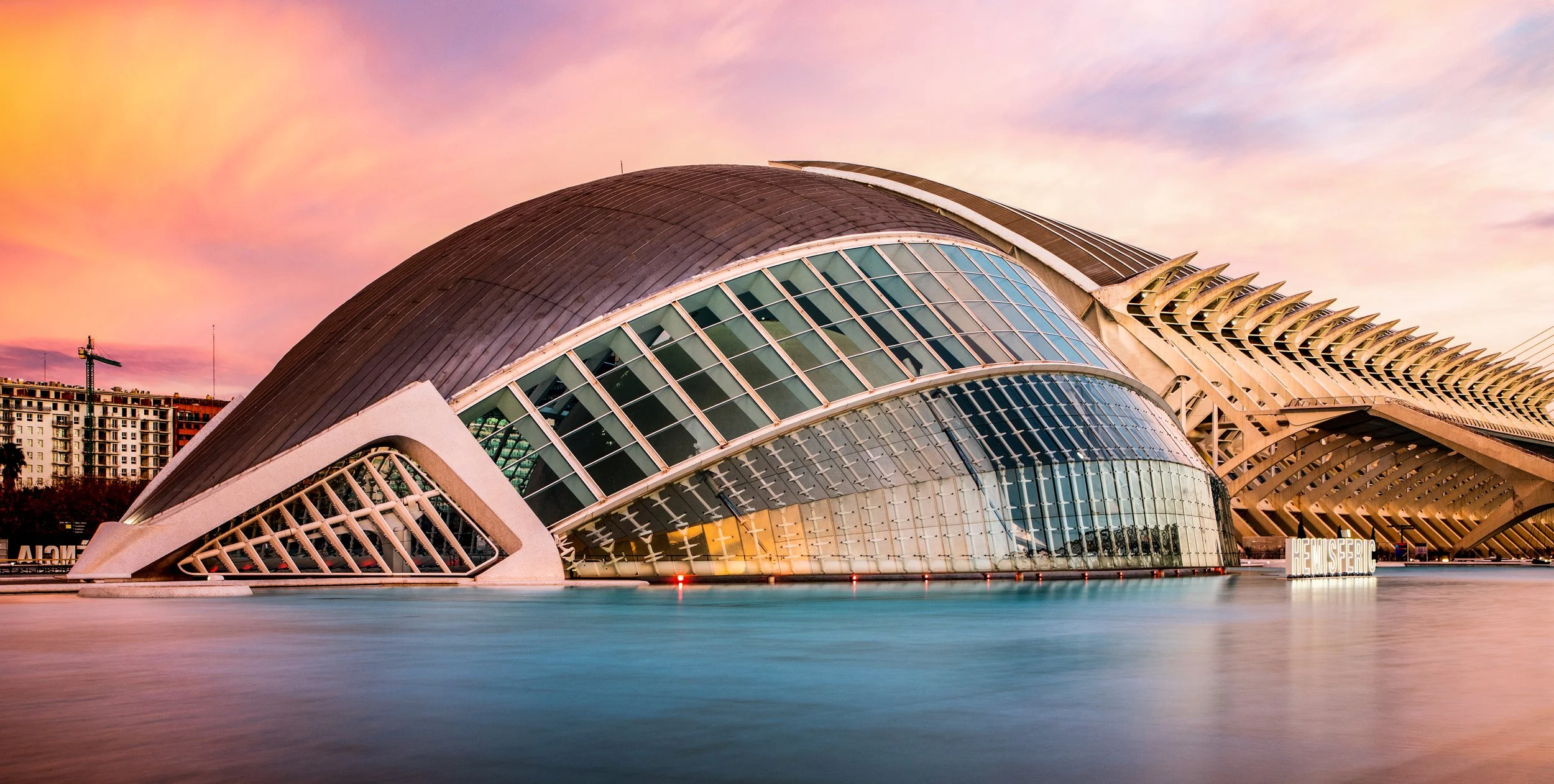 Modern architectural building with curved glass and metal structure, located near water with a colorful sky at sunset.