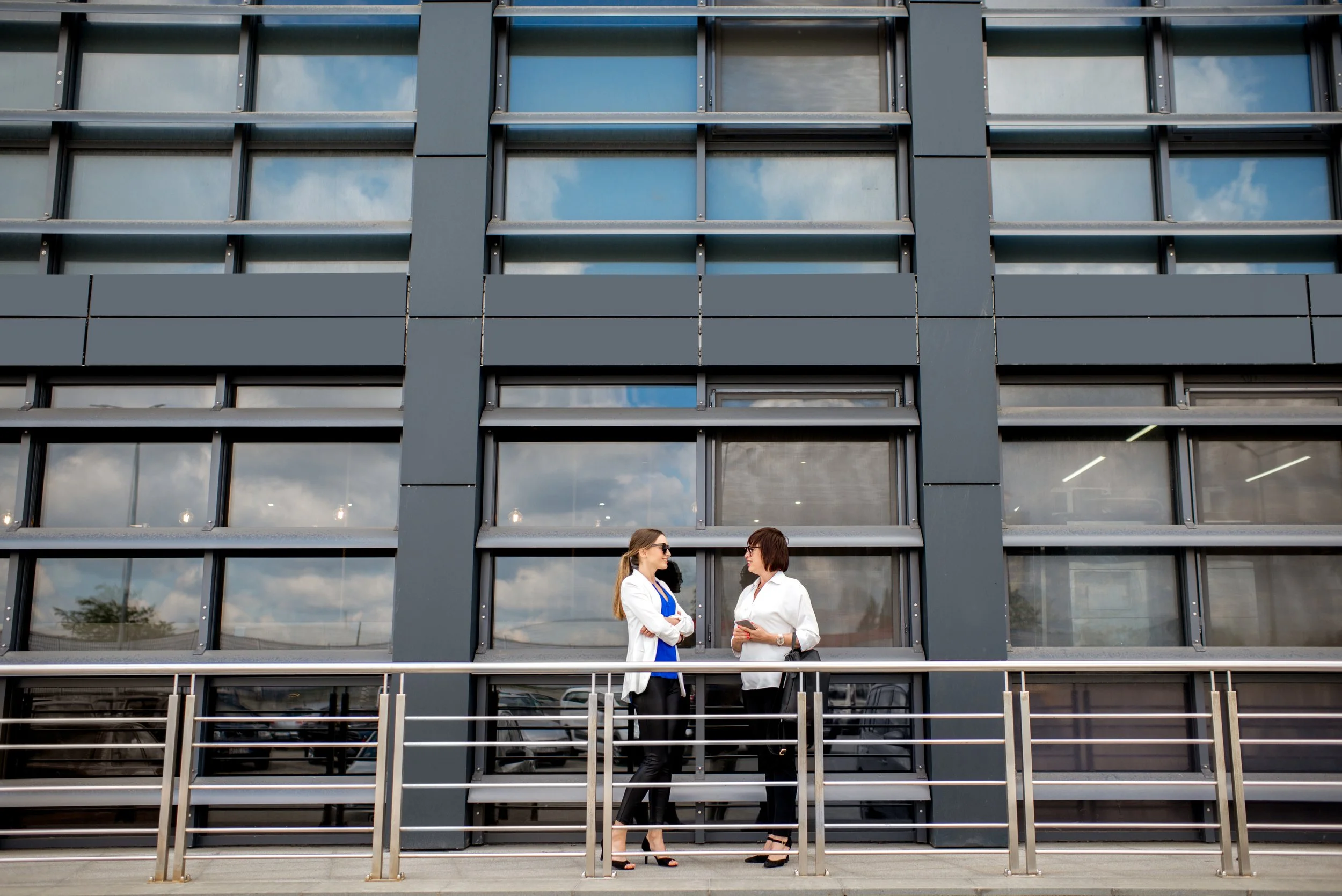 Two women in business attire talking outside a modern glass office building.