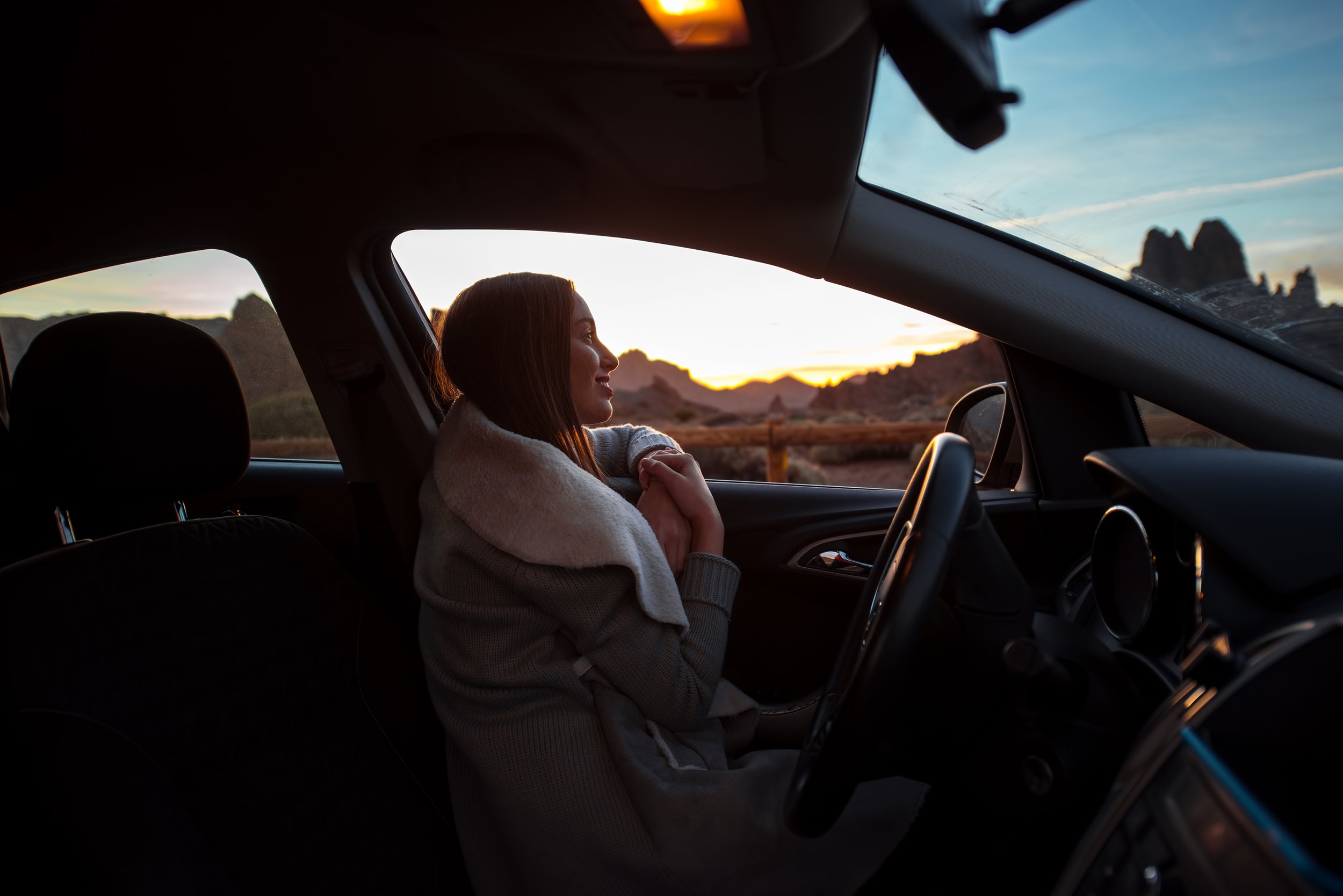 A woman sitting in the driver's seat of a car during sunset, looking out the window at a mountainous landscape with a blue sky.