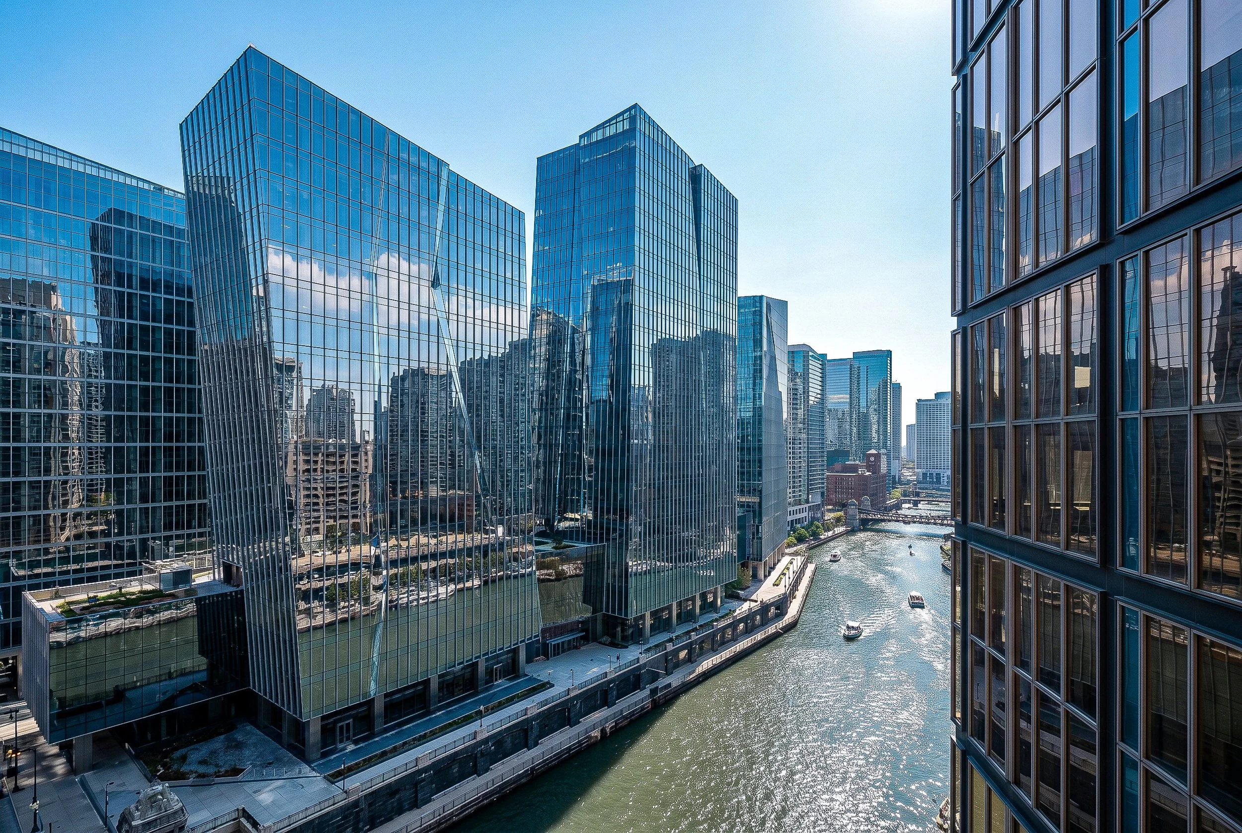 View of modern glass office buildings along a river with a bridge, city skyline, and clear blue sky in the background.