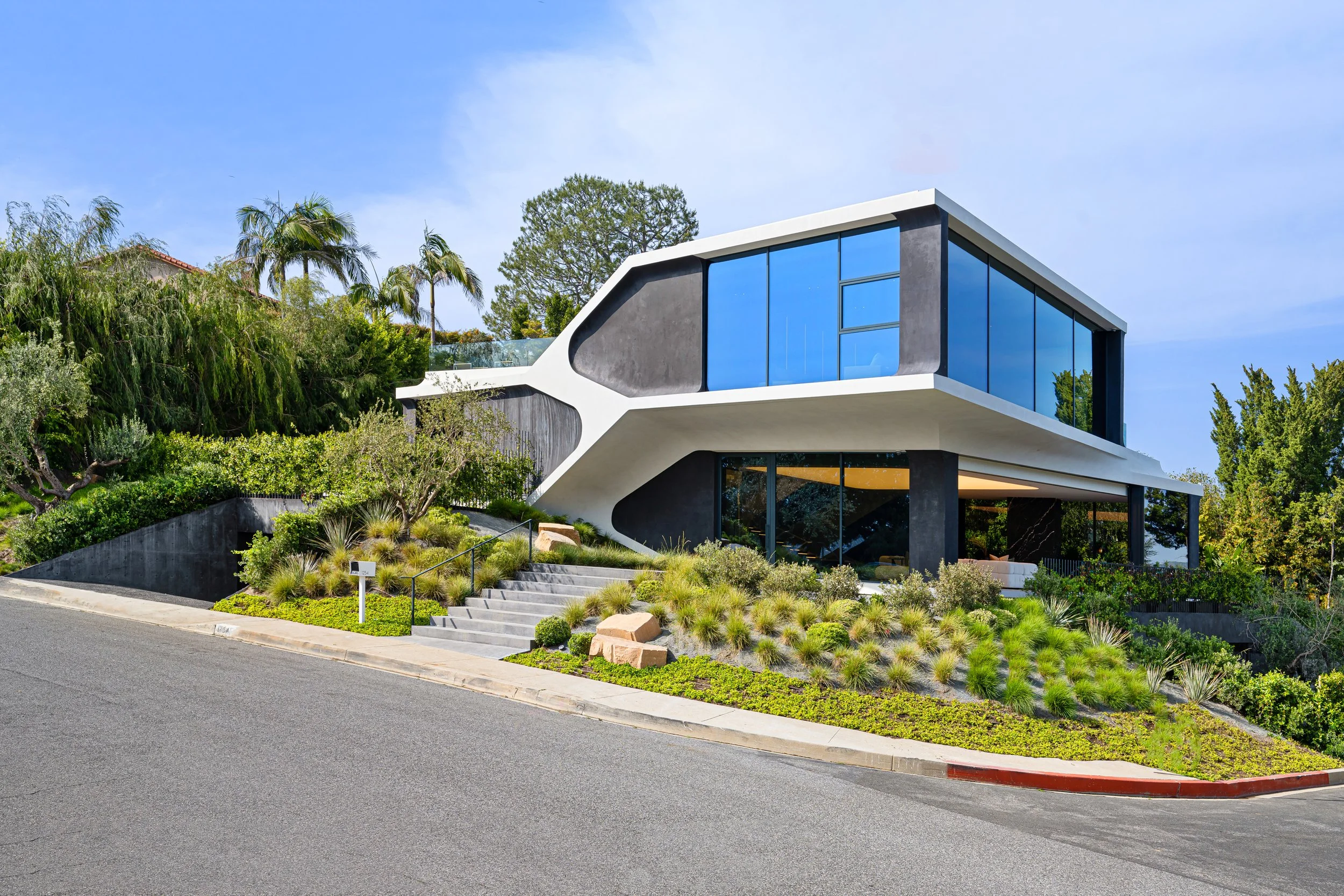 Modern house with large glass windows and an overhanging white roof, surrounded by lush greenery and a suburban street with steps leading up to it.