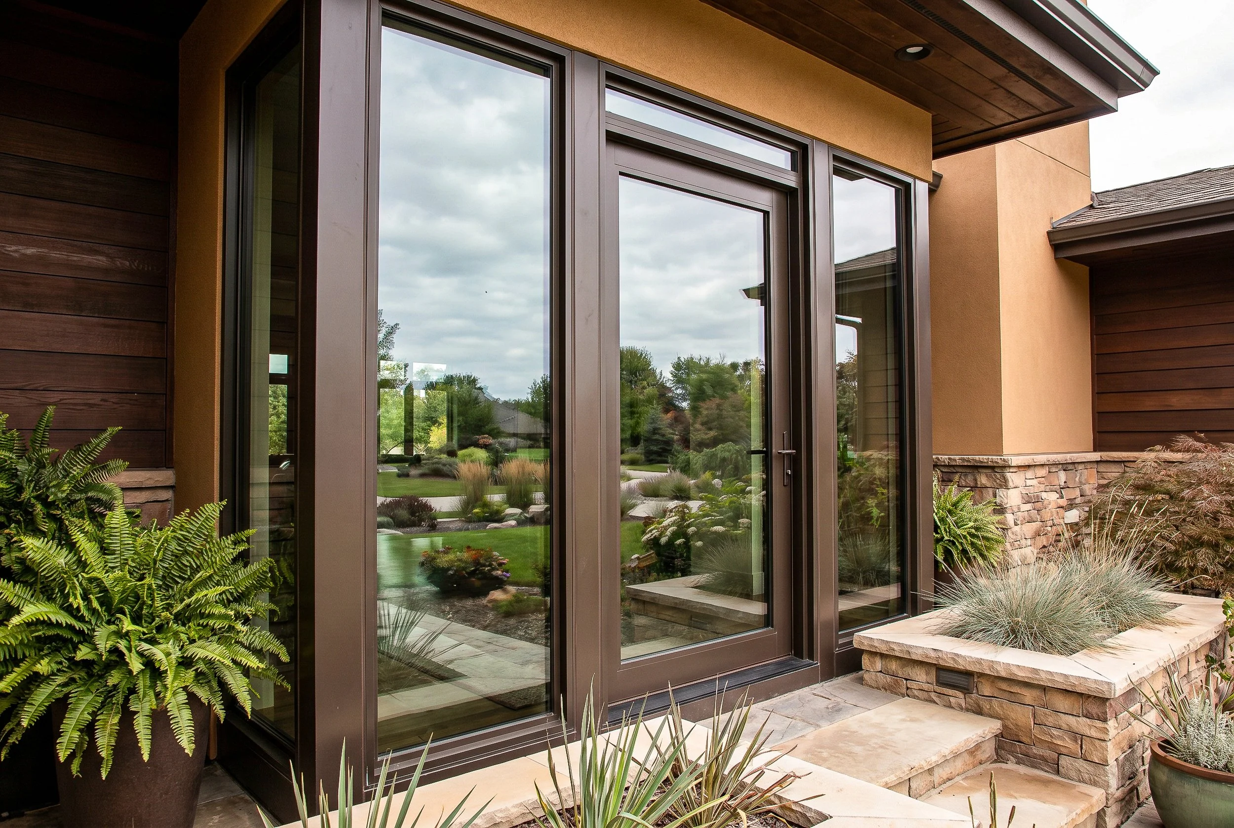 An exterior view of a modern house showing large glass sliding doors, a stone and stucco facade, surrounding greenery, and landscaped plants in pots.