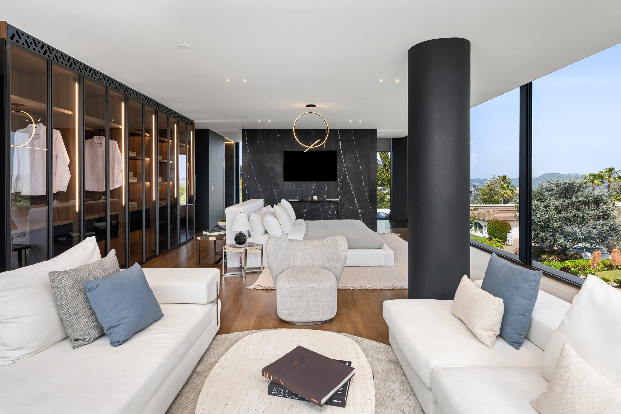 Modern living room with white sofas, gray pillows, round white rug, black marble fireplace wall, large windows with a view of trees and houses, and contemporary circular ceiling light.