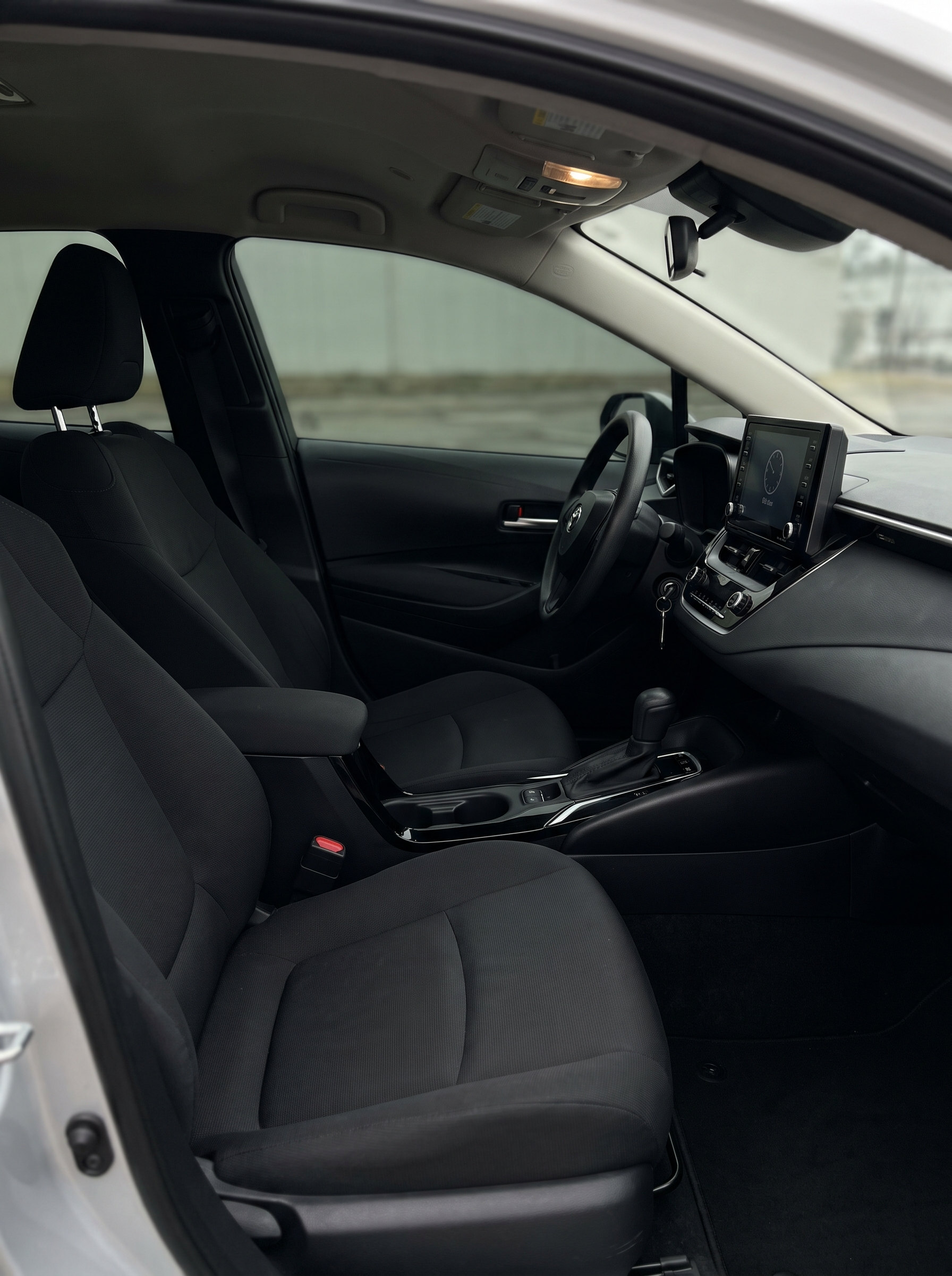 The interior of a car showing the front seats, dashboard, steering wheel, and center console with controls and a digital display. The driver and passenger seats are gray fabric, and the interior ceiling has lights and controls.