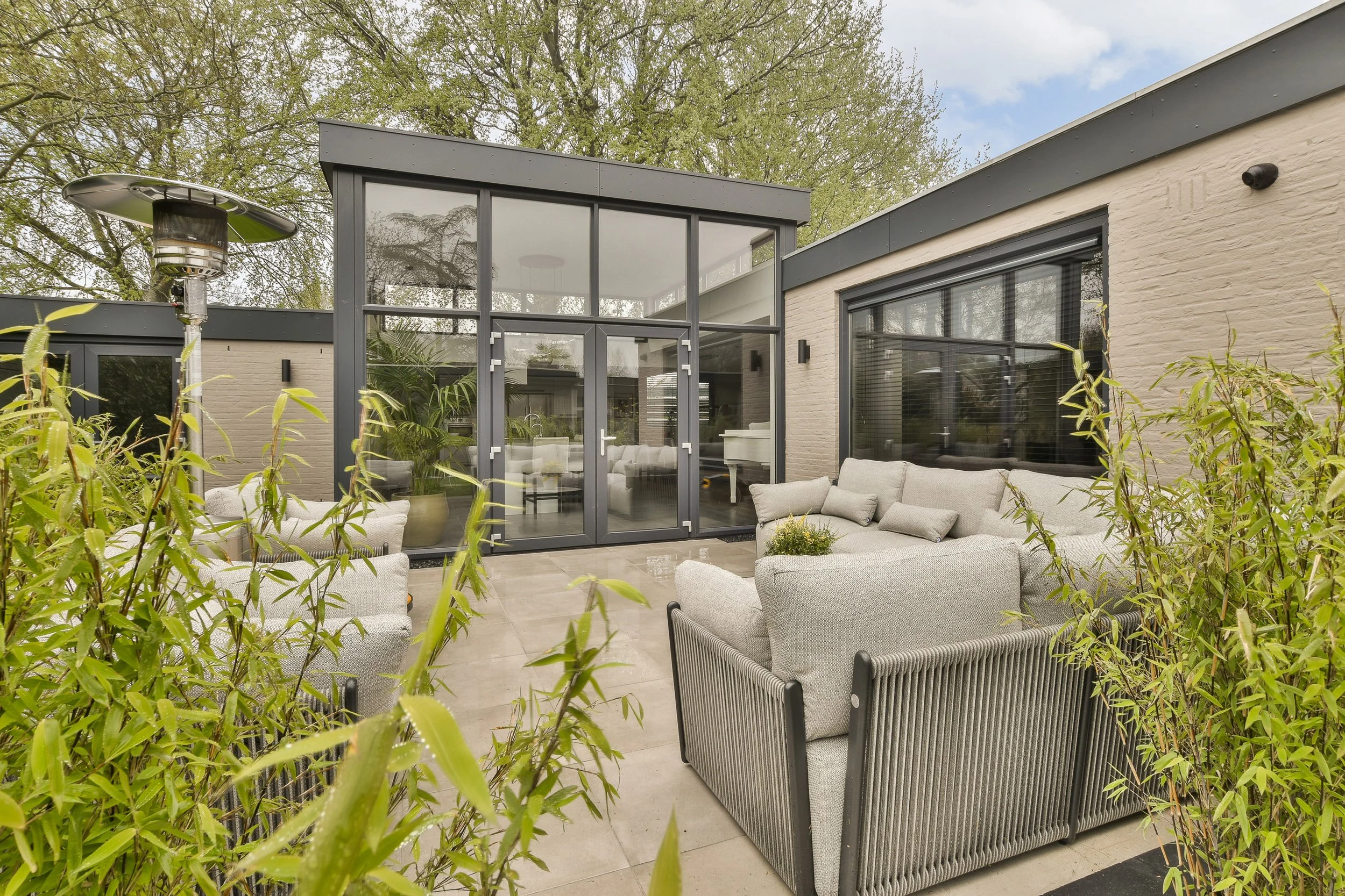 Outdoor patio with gray wicker furniture and beige cushions, surrounded by green plants, with a large glass door and windows leading to an indoor room, and a heater in the background. Trees with green leaves are visible in the sky.