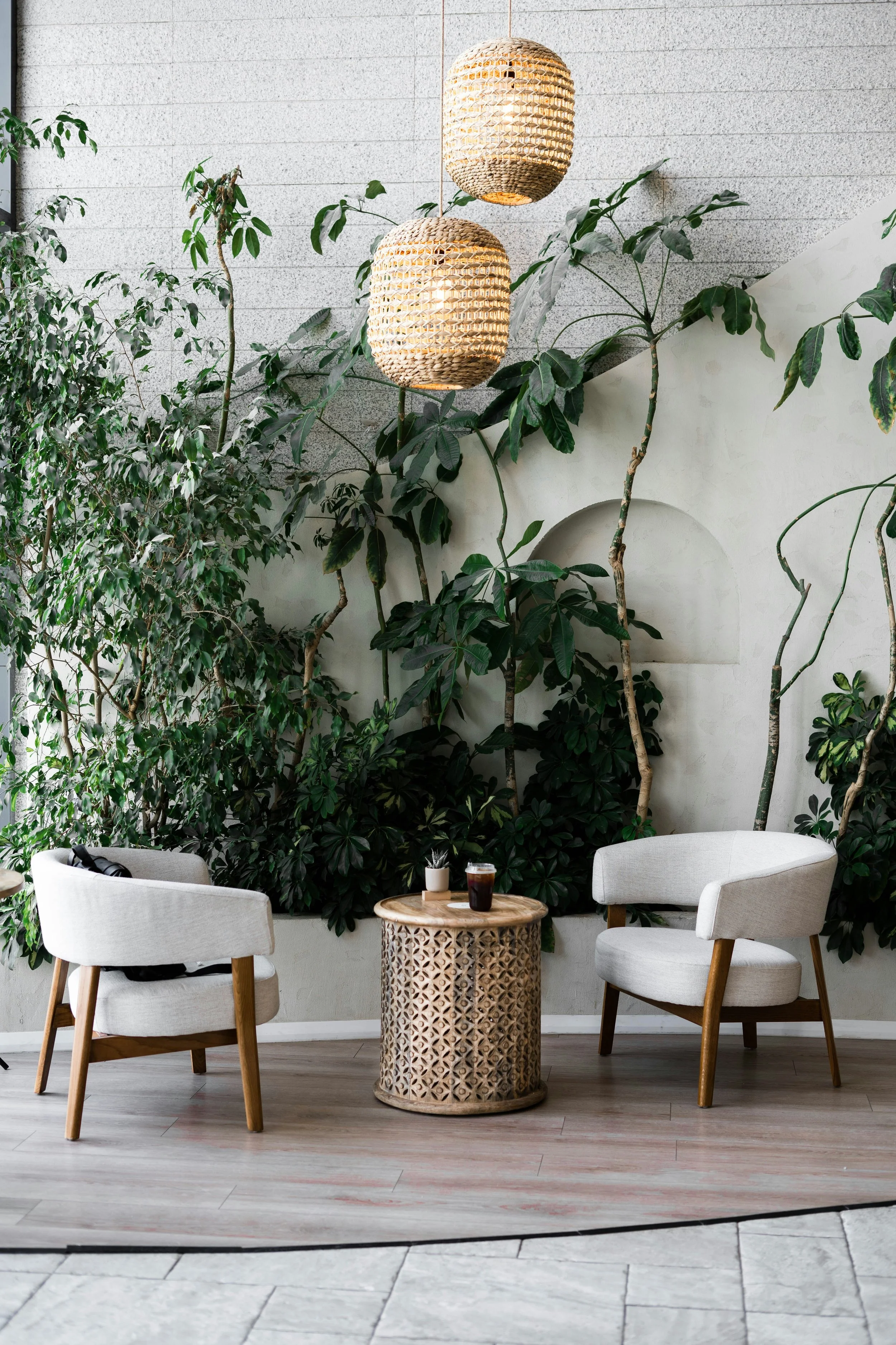 Two white armchairs with wooden legs are positioned on either side of a small, round wooden lattice-patterned table. On the table are a small potted plant, a glass of dark-colored beverage, and a smaller dark-colored container. Behind the chairs, lush green plants and trees grow along a white wall with textured bricks, topped by two wicker pendant lights hanging from the ceiling.