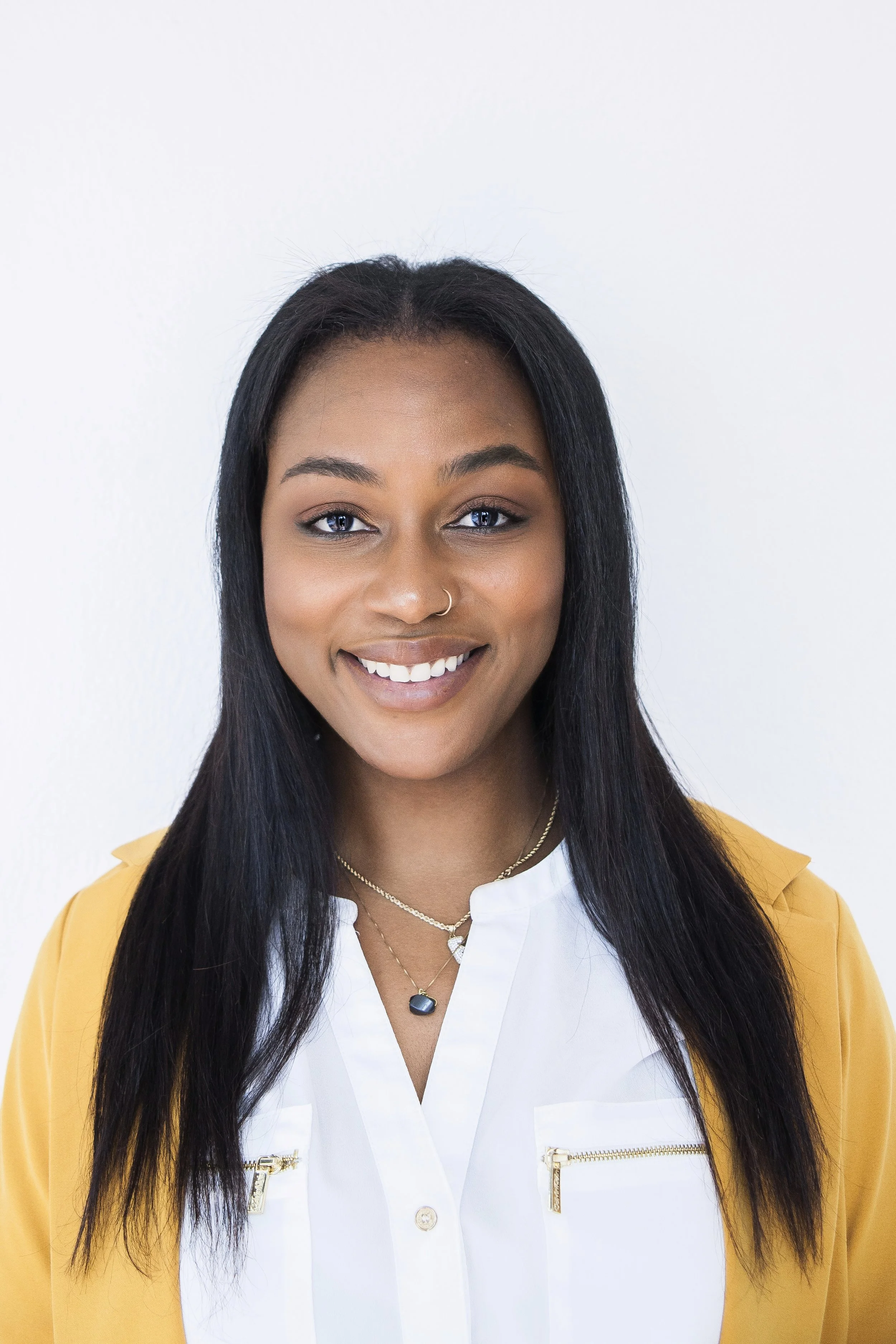 A smiling Black woman with long black hair, wearing a yellow blazer over a white blouse, and layered necklaces, standing against a white background.