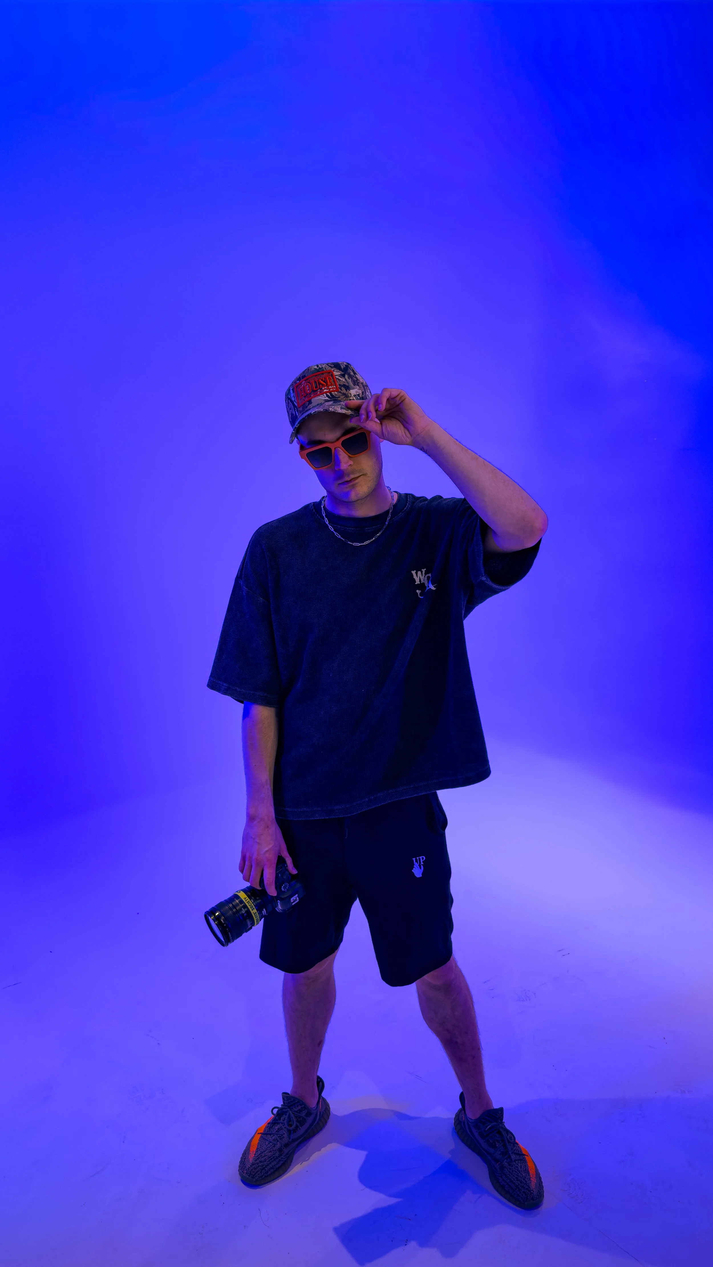 A young man in casual streetwear stands against a blue-lit background, wearing a camo baseball cap, orange sunglasses, a black t-shirt, black shorts, sneakers, and holding a camera.