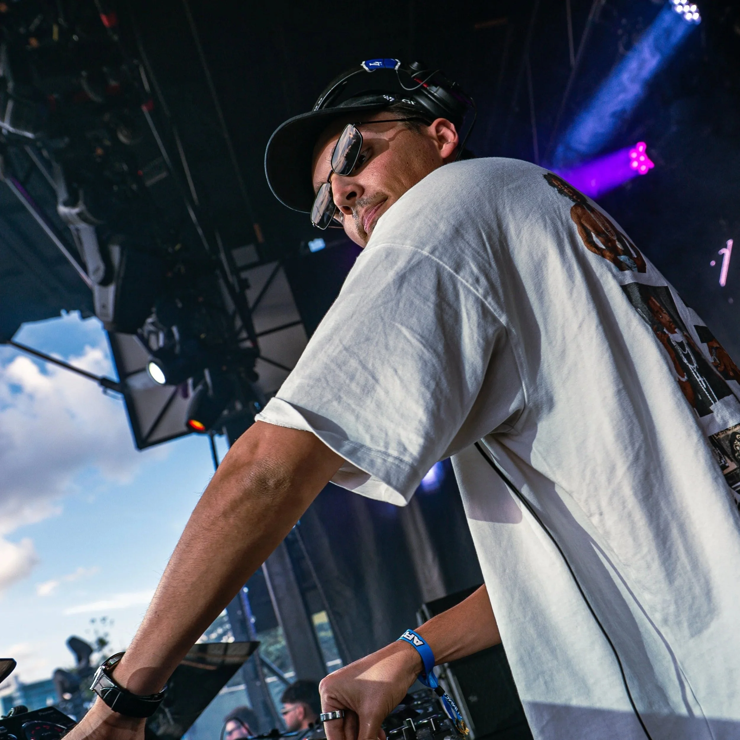 A DJ wearing sunglasses, a backwards cap, and a white t-shirt at an outdoor event, operating a DJ mixing console under a tent, with a bright blue sky and clouds in the background.