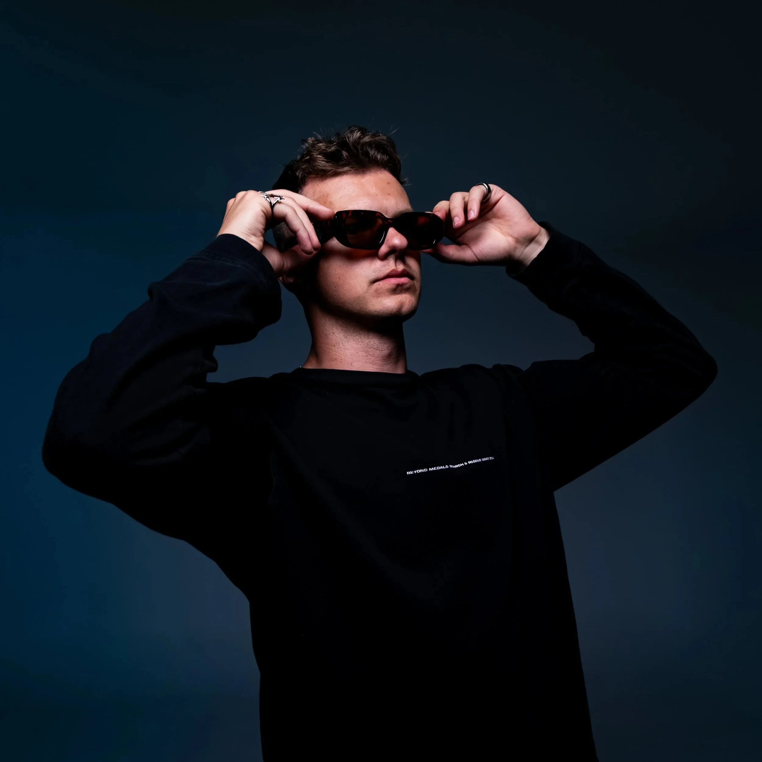 Person adjusting sunglasses against a dark background, wearing a black sweatshirt.