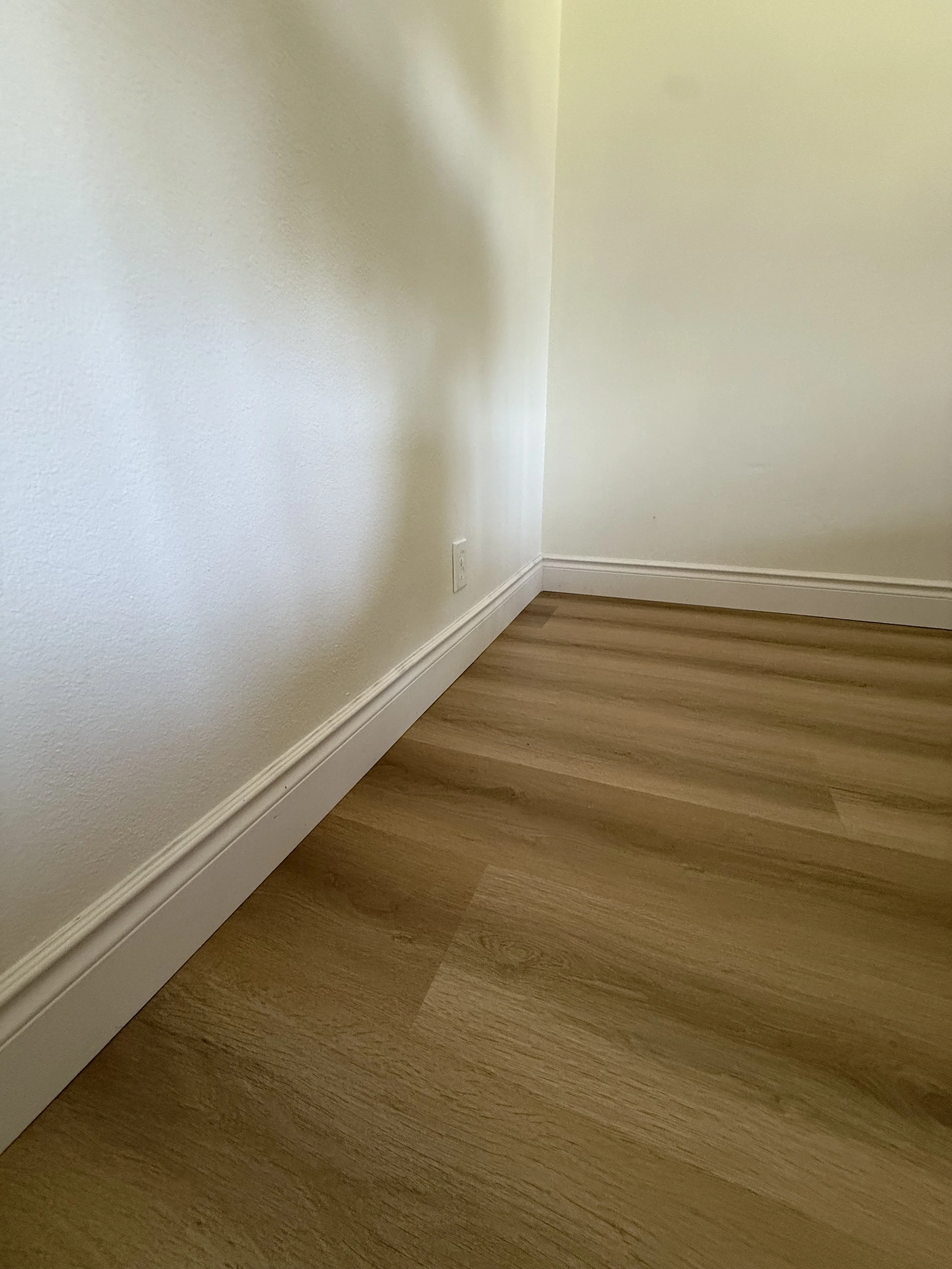 Corner of a room with white walls and wooden flooring, featuring baseboard molding along the bottom of the wall and an electrical outlet.