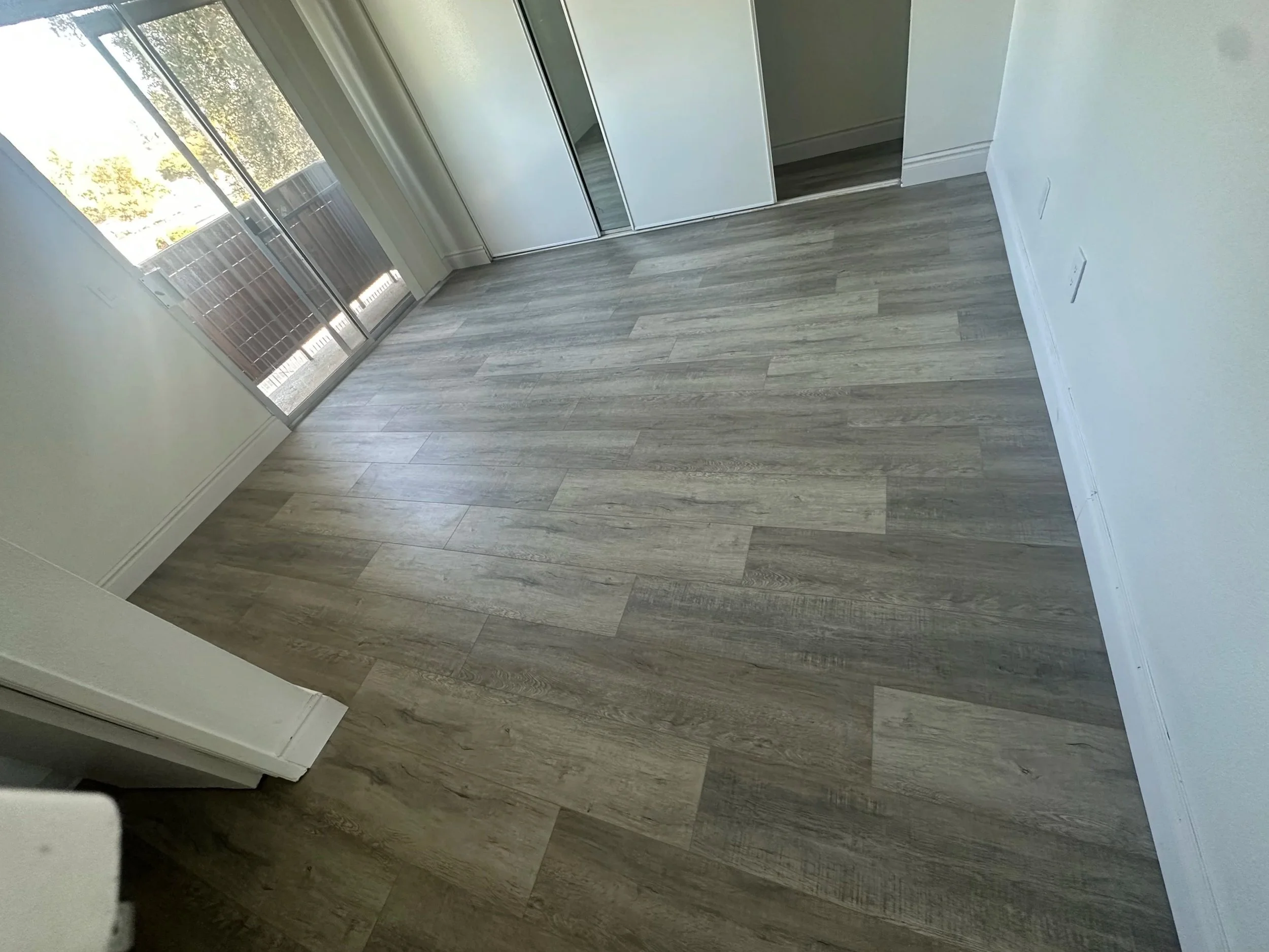 A room with light-colored wooden flooring, sliding glass doors leading to an outdoor patio with a railing, and a closet with mirrored sliding doors. Sunlight is coming through the glass doors, illuminating the room.
