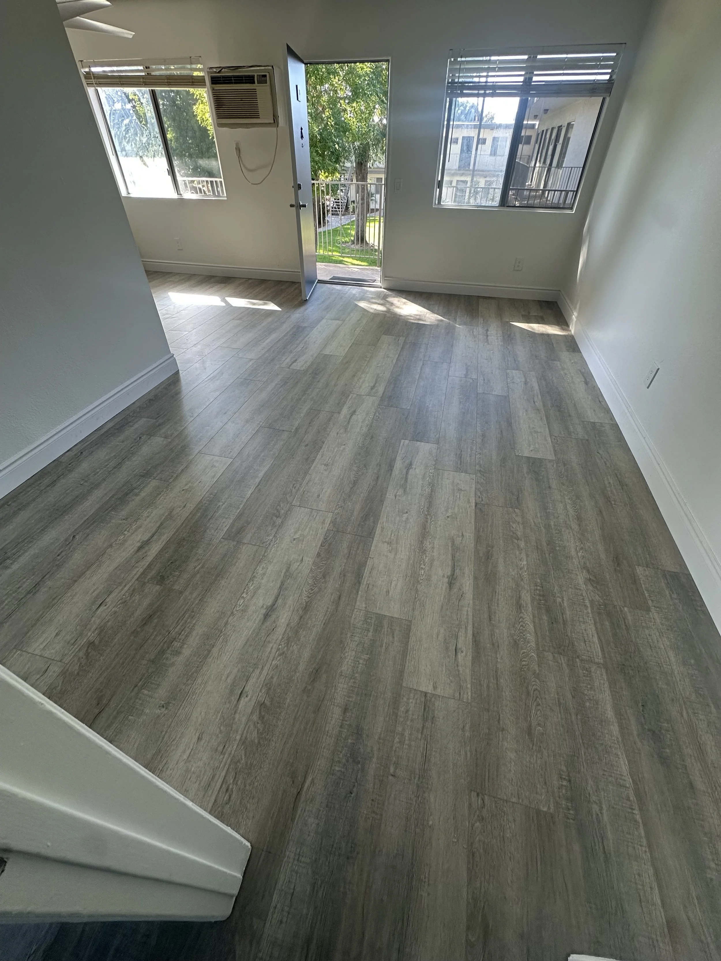 Empty room with wood-look flooring, white walls, windows showing outside view, and a door open to an outside balcony with trees.