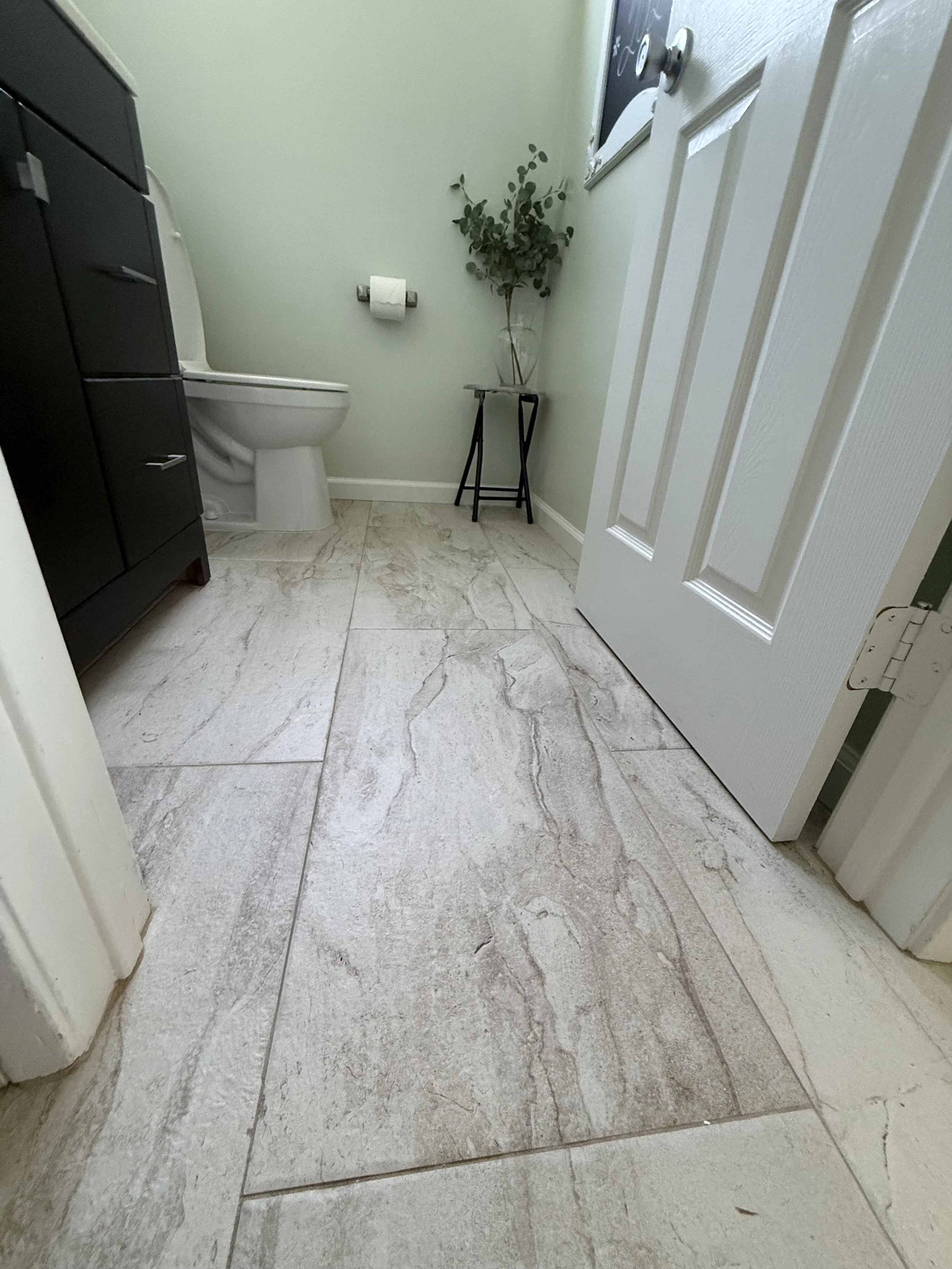 A bathroom with beige marble tile flooring, a white toilet, a dark cabinet on the left, a small black table with a glass vase holding green foliage, a toilet paper roll on the wall, and a partially open white door.