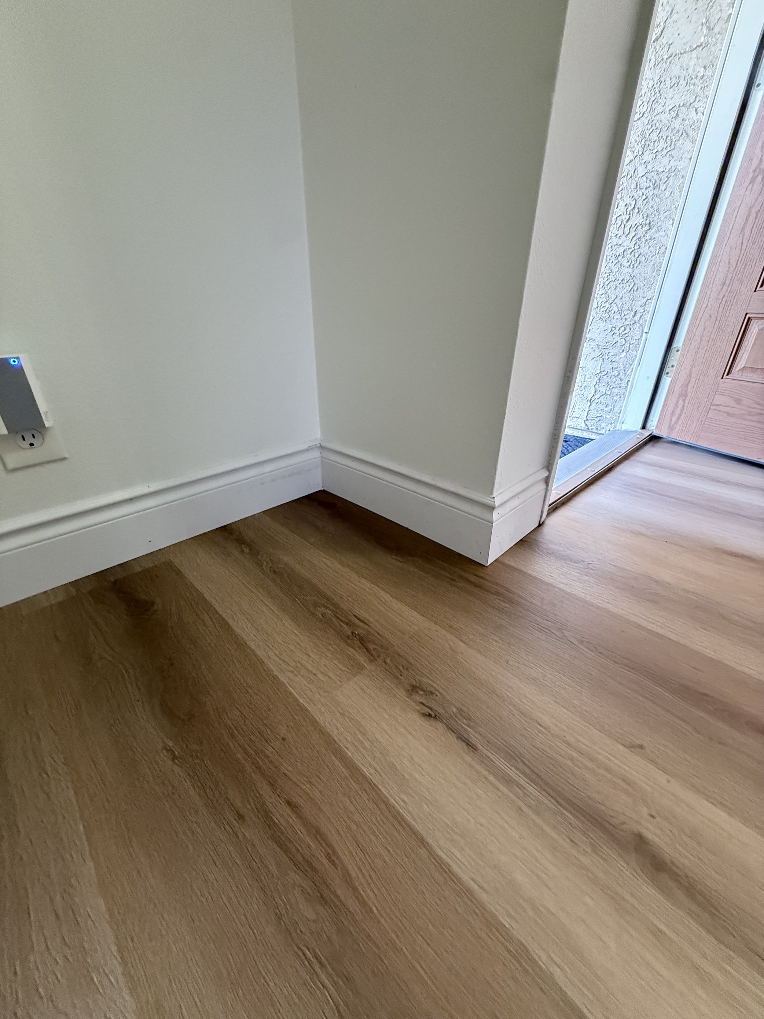 The image shows a corner of a room with light-colored walls, a wooden floor, a baseboard, an electrical outlet, and a doorway leading outside with a wooden door and textured exterior wall.