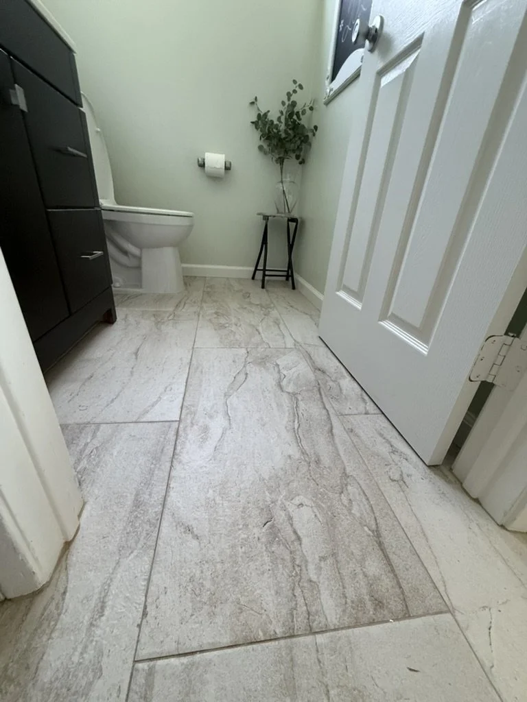 Bathroom with gray marble tile floor, a toilet, black cabinet, small stand with green plant, closed white door, and laundry appliances.