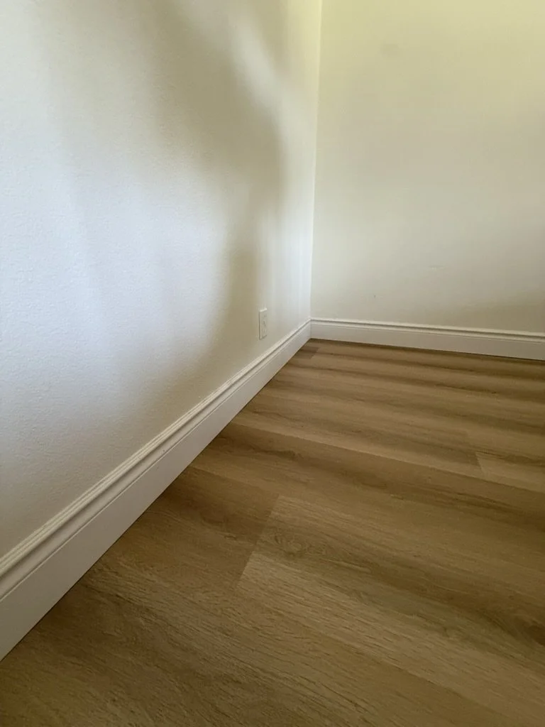 Empty corner of a room with white walls, a power outlet, wooden flooring, and white baseboards.