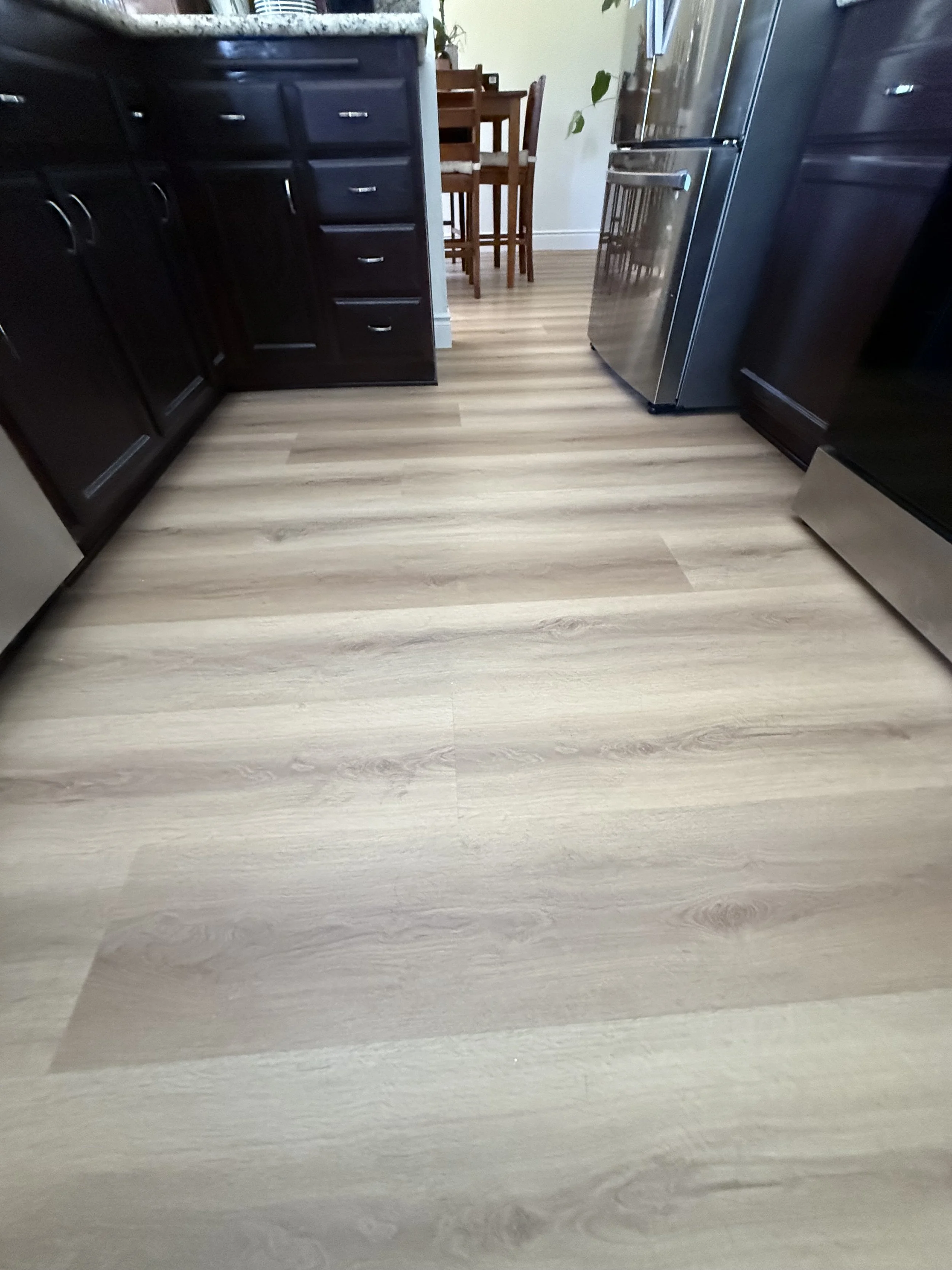 A kitchen with dark cabinets, a stainless steel refrigerator, a wooden dining table with chairs, and light-colored wood flooring.