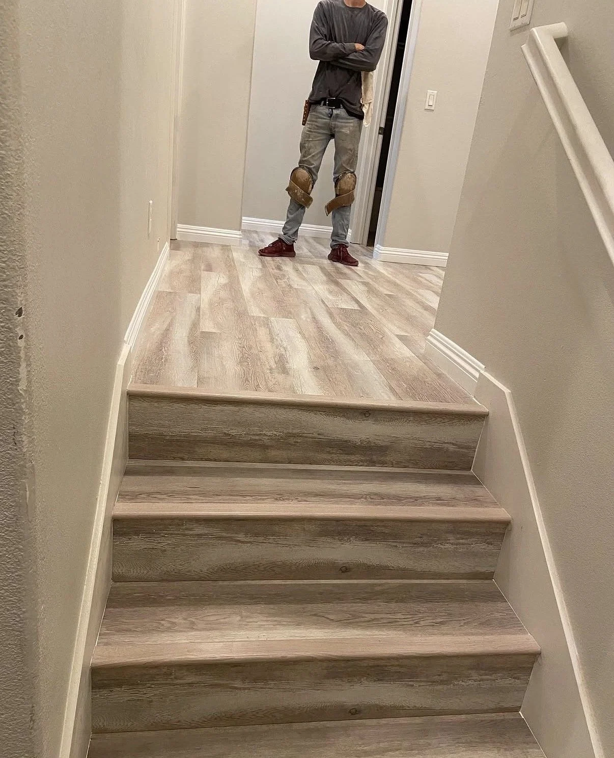 Photo of a person standing at the top of wooden stairs, wearing gray pants, a dark top, and red shoes, with their arms crossed, inside a house hallway with beige walls and wood flooring.