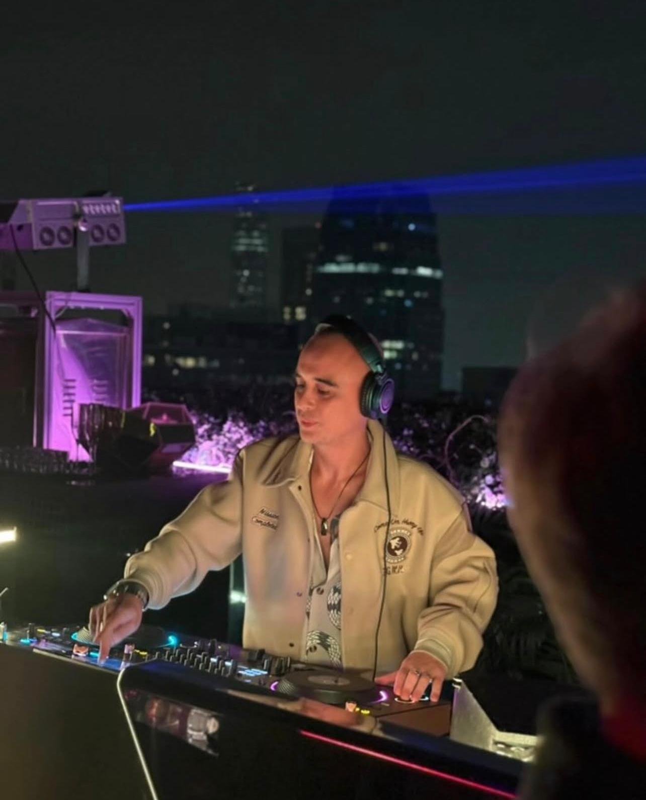 DJ Craig Lopez. A male DJ wearing headphones and a beige jacket, playing music at a rooftop party during nighttime with Hong Kong city skyscrapers in the background, illuminated by purple and blue lights.