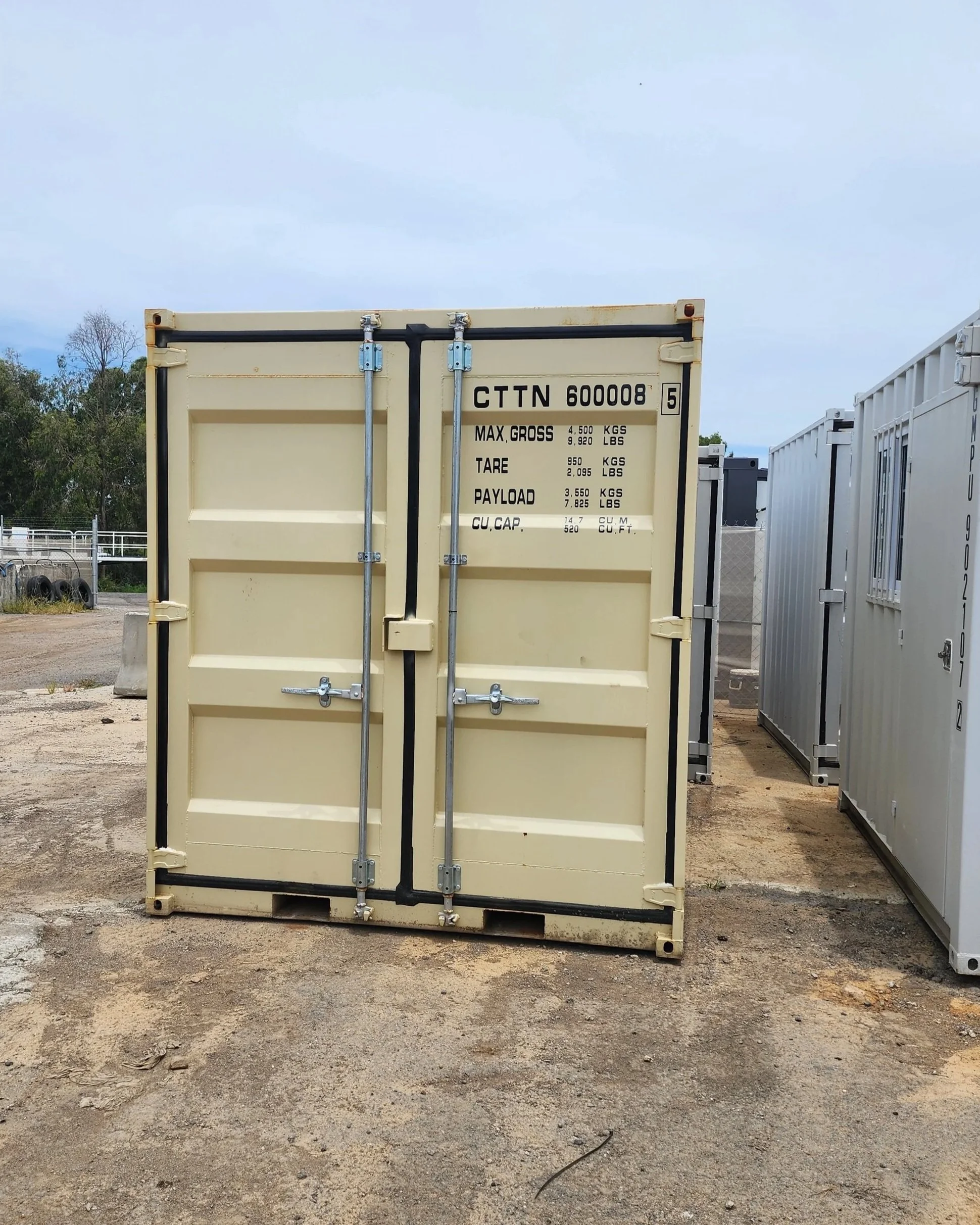Front view of a beige 20-foot shipping container with locking doors and capacity markings, next to white containers in an outdoor storage yard