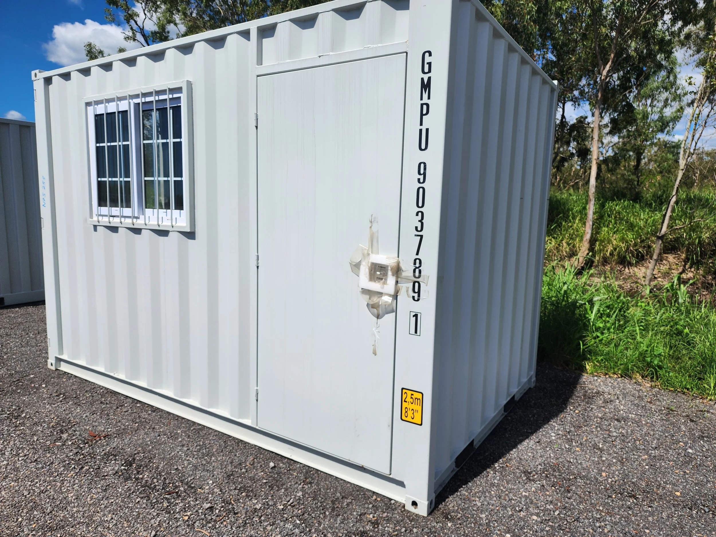 White shipping container with barred window and locked doors outdoors near trees