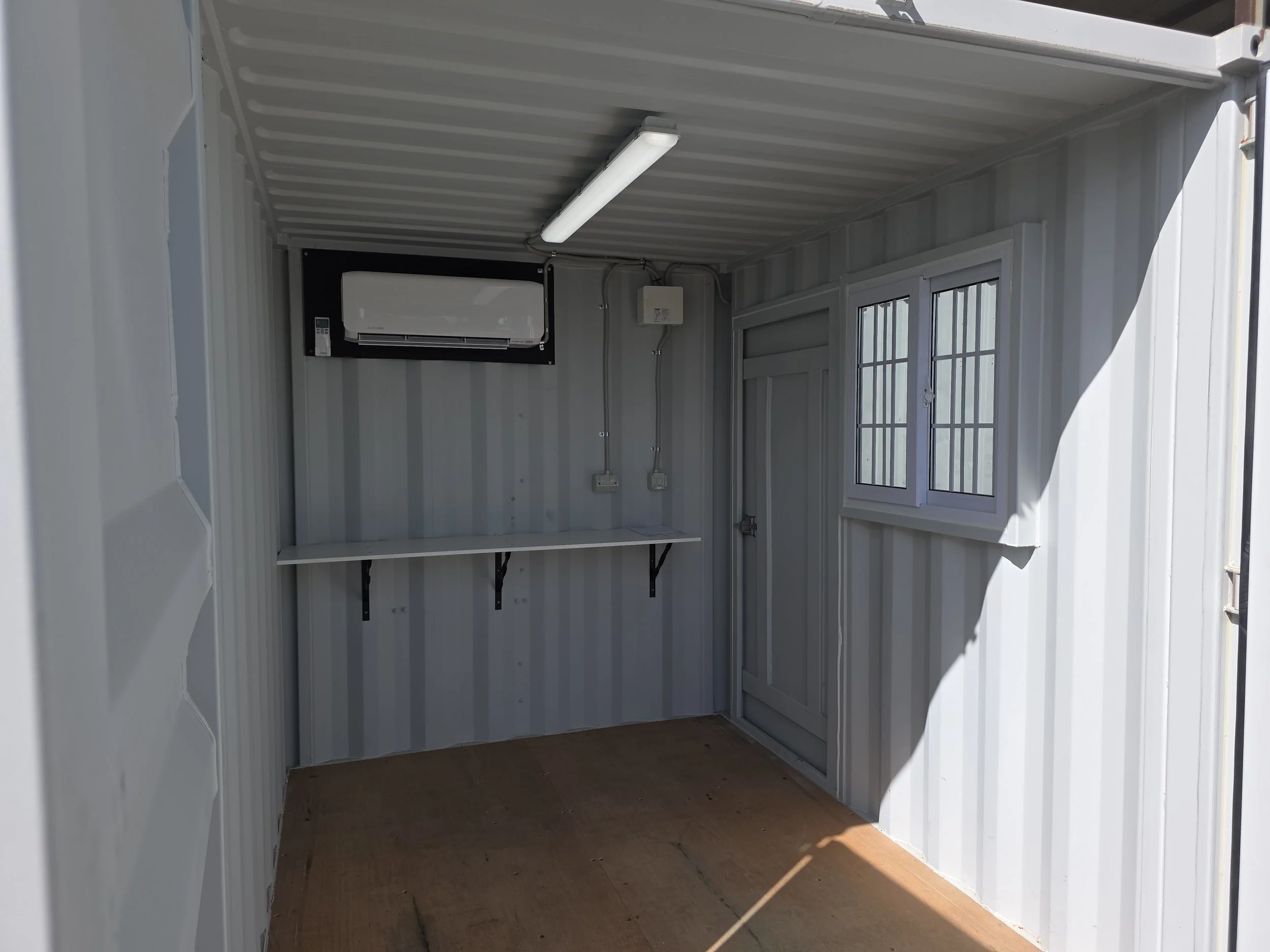 Interior of a converted shipping container office with air conditioner, fluorescent ceiling light, workbench, door, and barred windows.