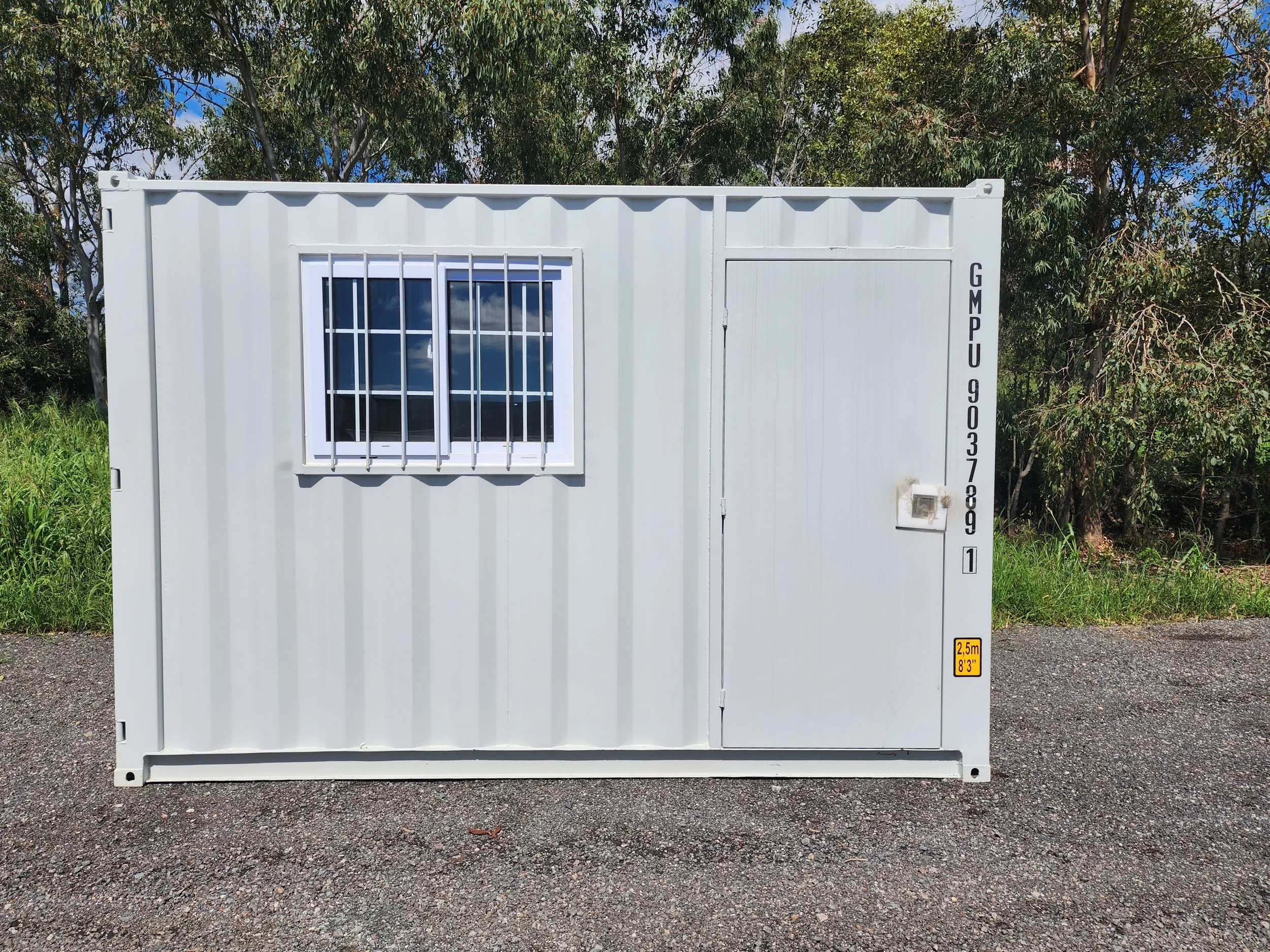 White modified shipping container with barred window, door, and height warning label outdoors among trees