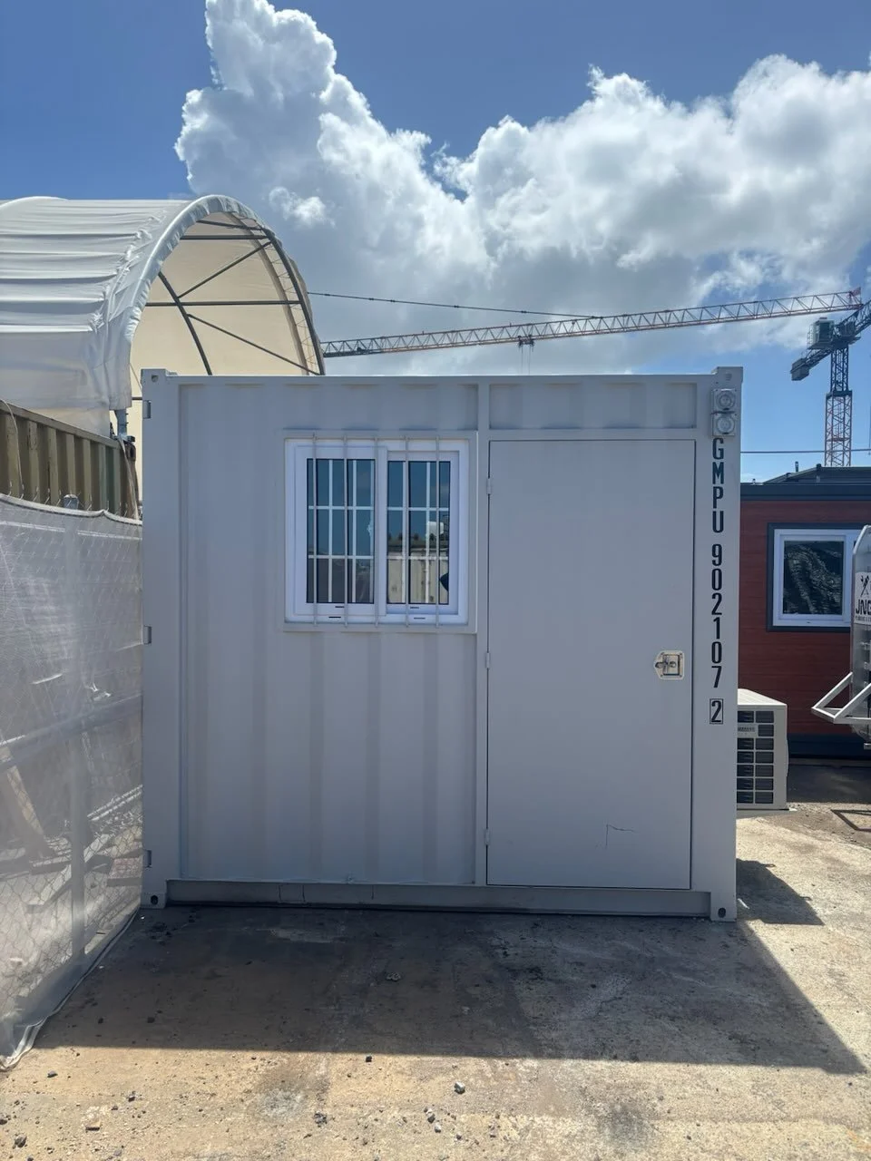 White portable construction office with barred window and door beside a covered structure and tower crane under a blue sky