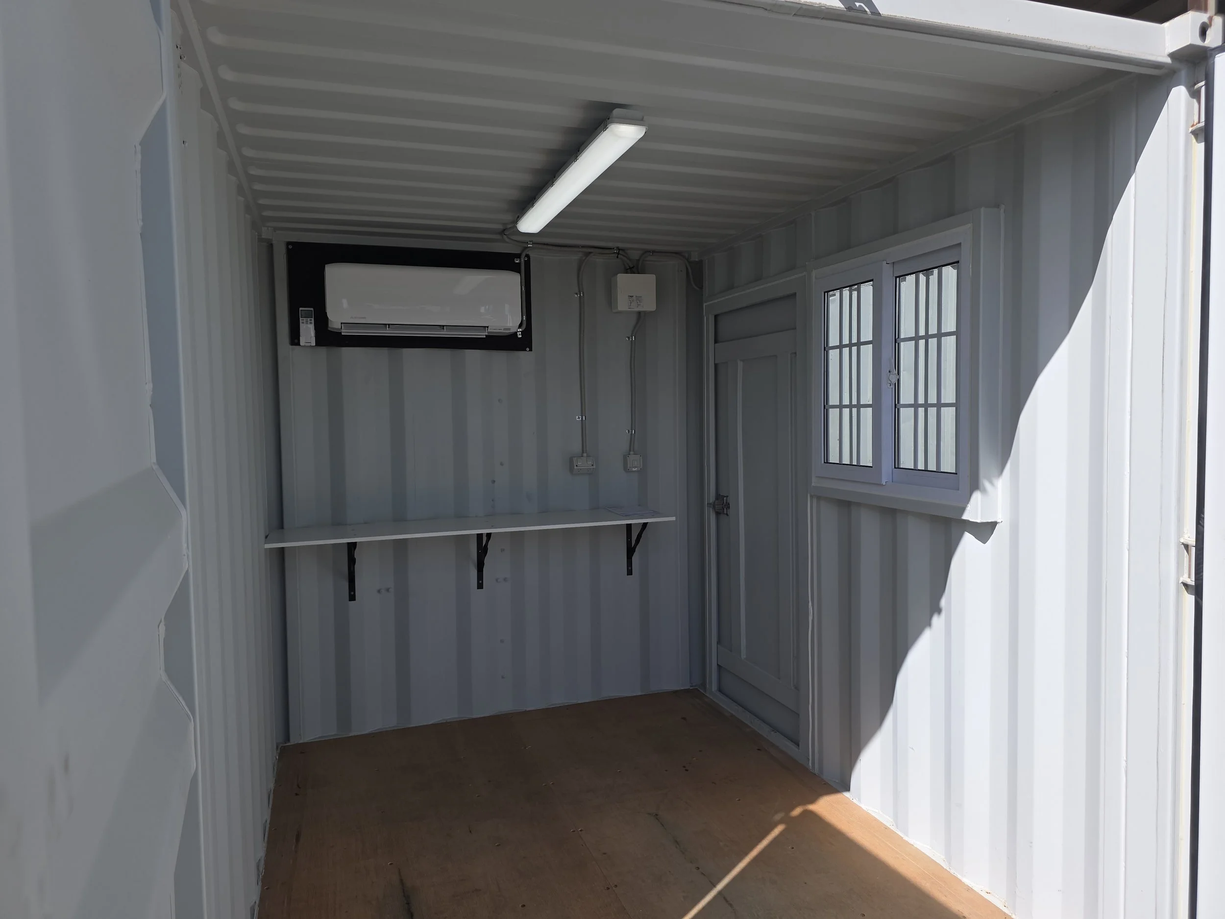 Interior of a converted shipping container with air conditioning, fluorescent lighting, workbench, electrical outlets, door, and barred window