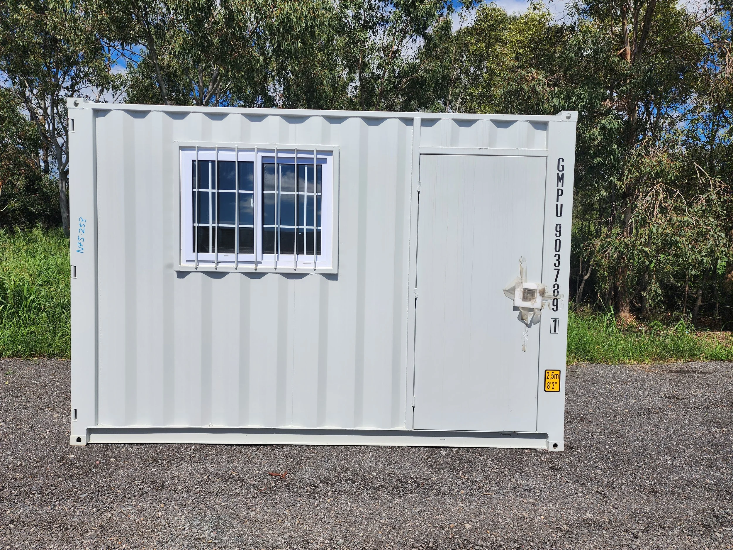 White converted shipping container with barred window, door, and exterior light on a gravel surface surrounded by trees