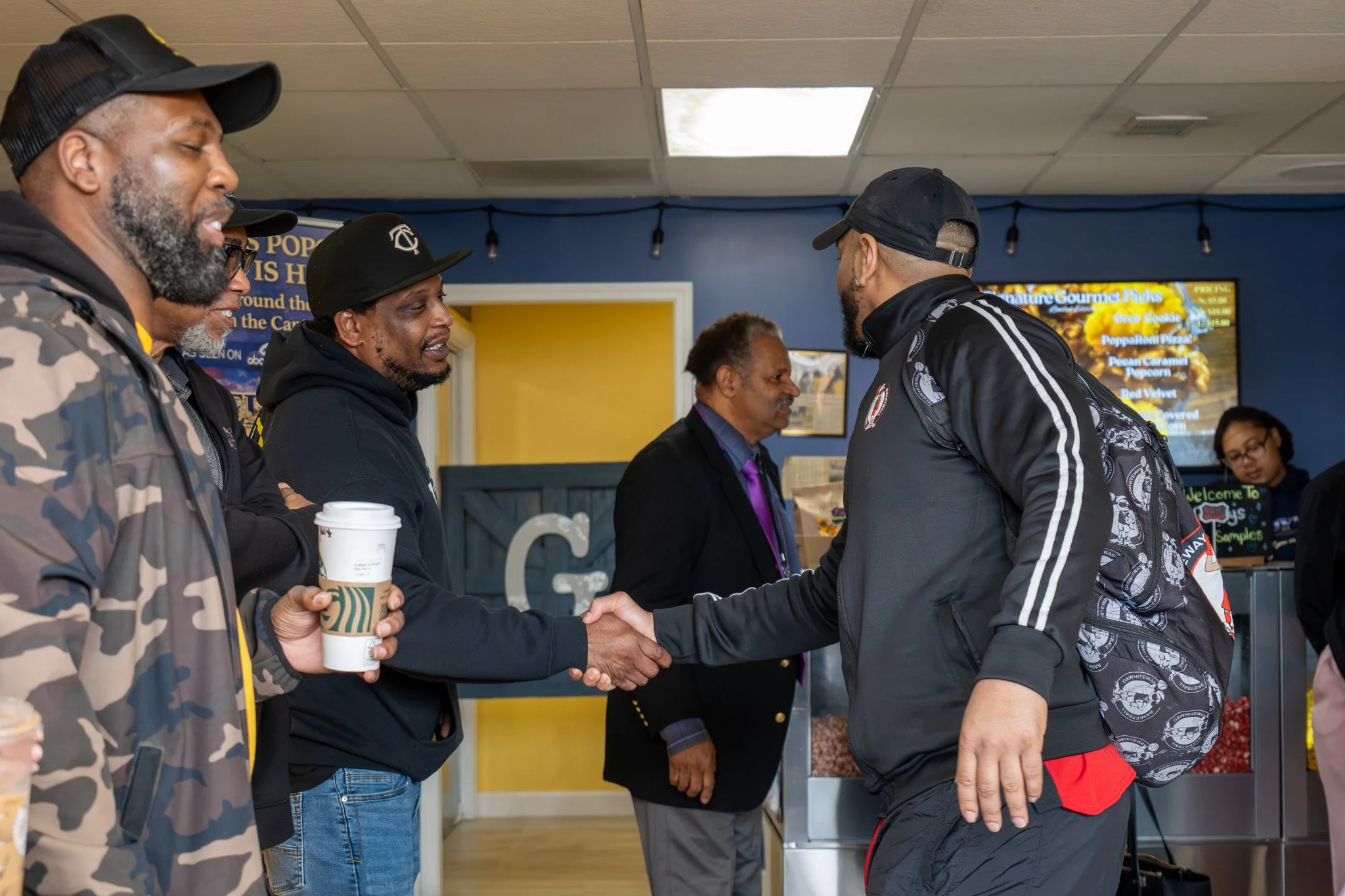 Four men shaking hands inside a cafe with a yellow and blue wall, with one man holding a Starbucks coffee cup, and a woman at a counter in the background.