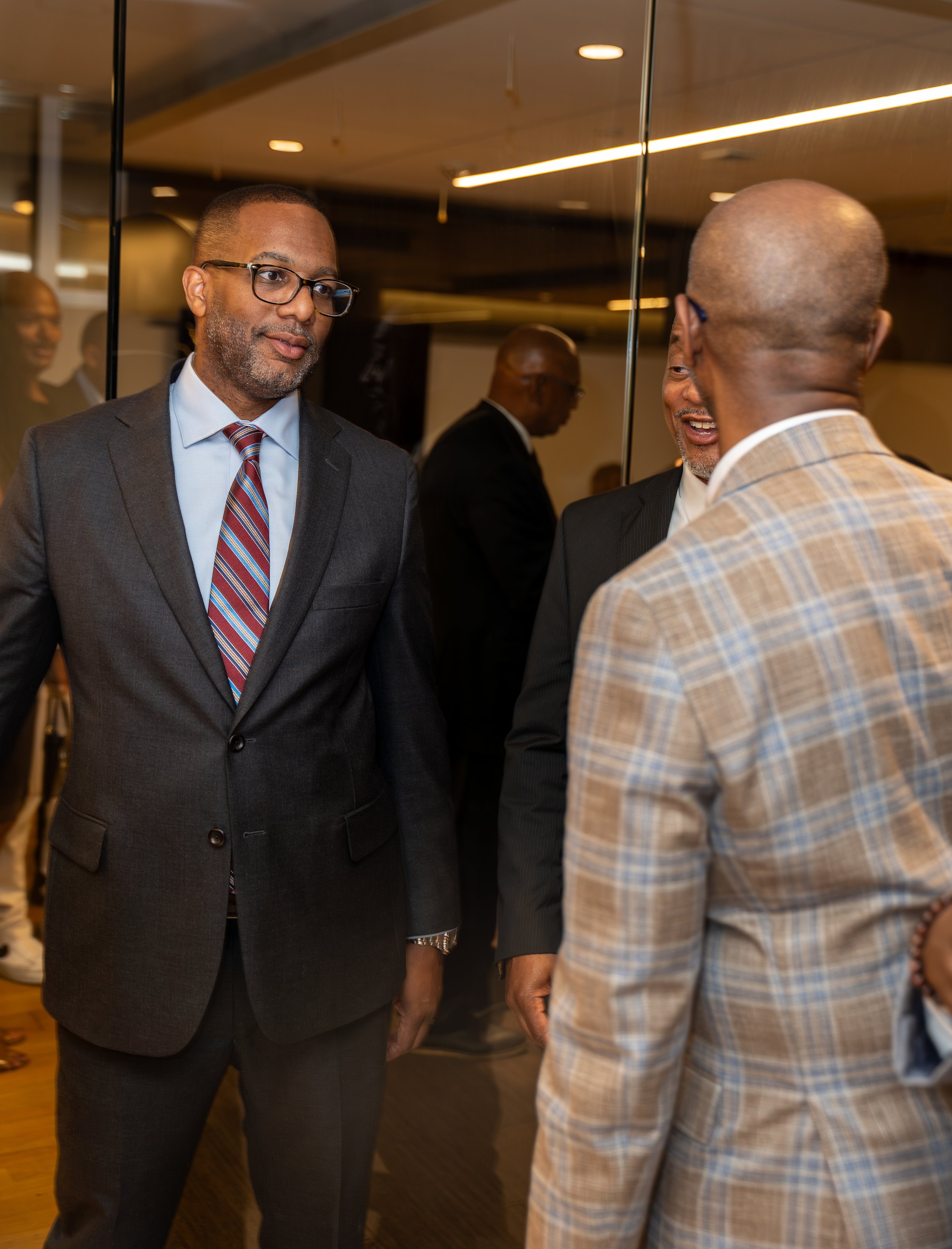 Three men in suits engaged in conversation at a professional event, with a glass wall and other people in the background.