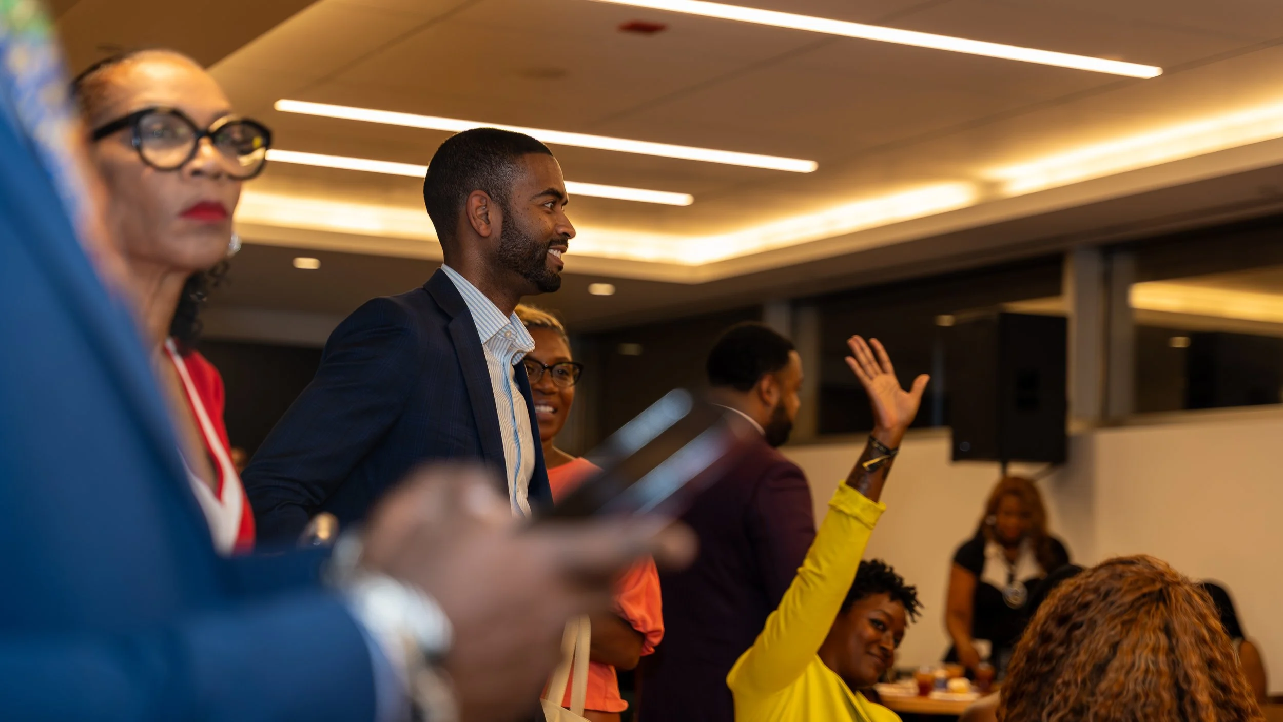 People attending a professional event or conference, some engaging and listening, with a woman in a yellow blazer raising her hand.