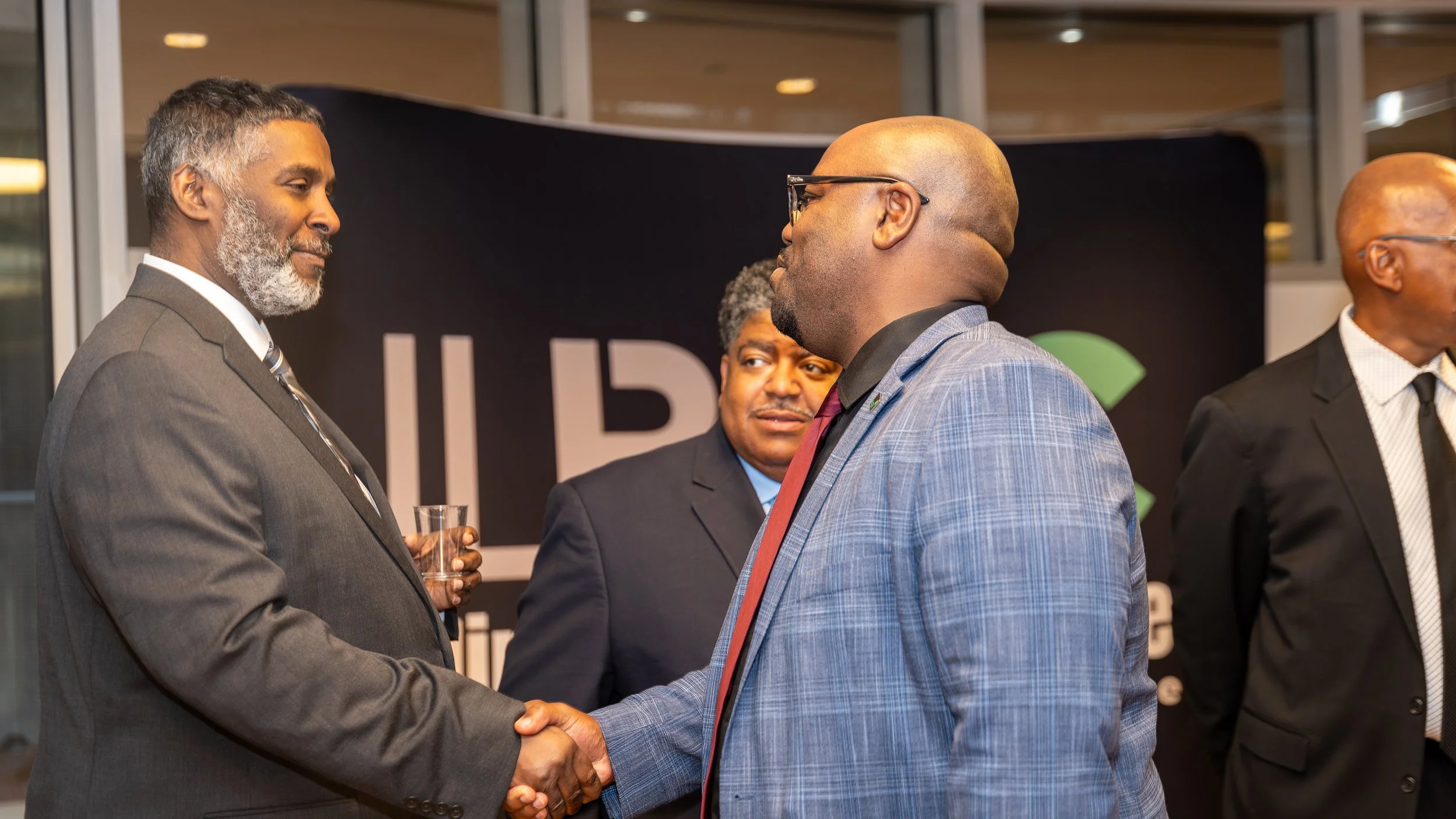 Two men in suits shake hands at a professional event, with a third man observing. The background features a black banner with partially visible white and green text.