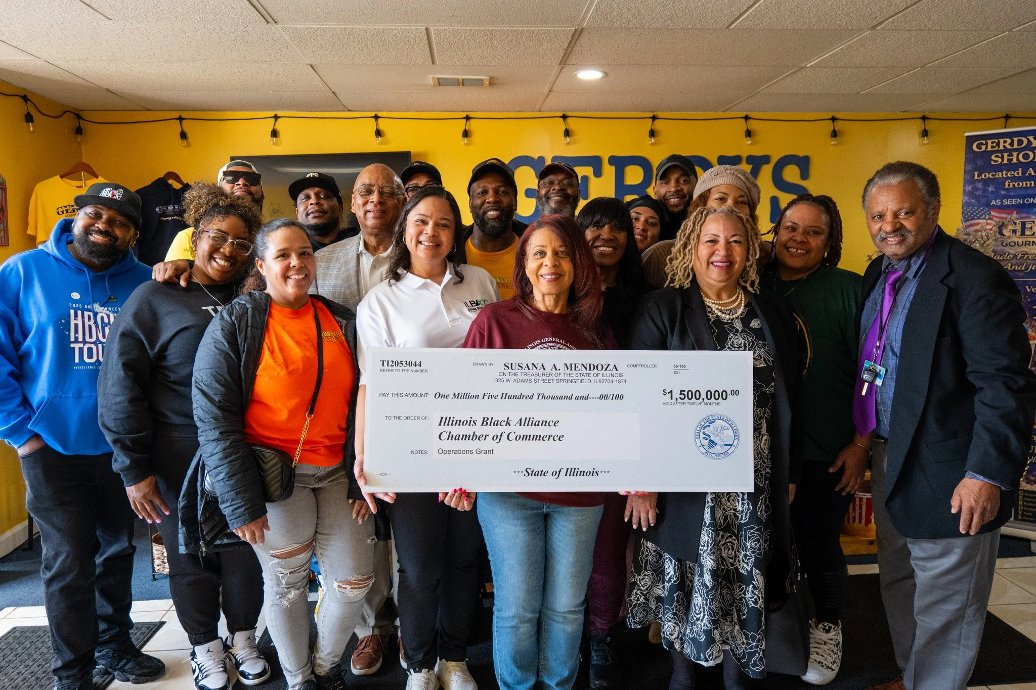 Group of diverse people standing together indoors, holding an oversized check for one million five hundred thousand dollars made out to the Illinois Black Alliance Chamber of Commerce, with a yellow wall and various signs and clothing items in the background.