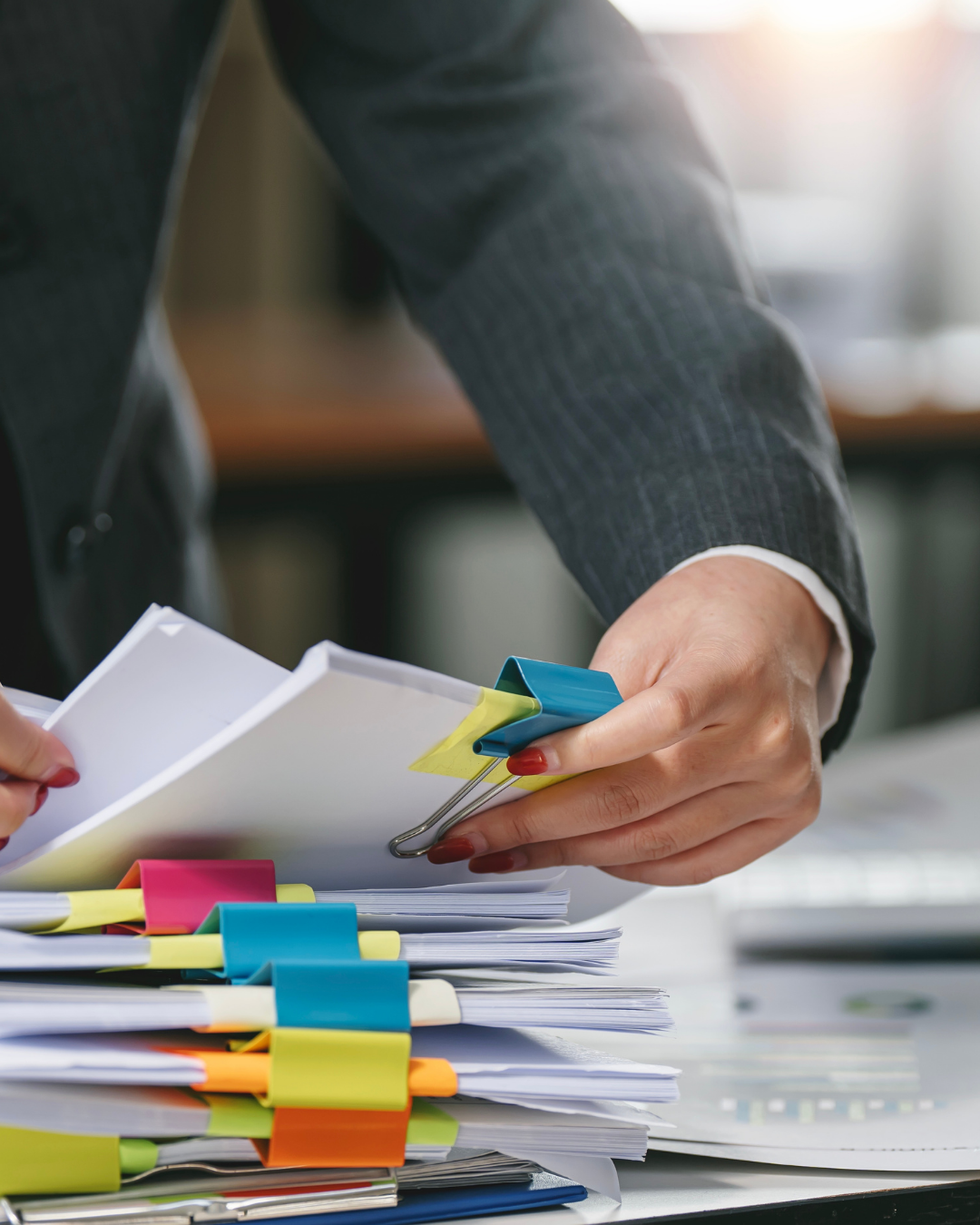 Person in a suit organizing and managing folders, papers, and sticky notes on a cluttered desk.