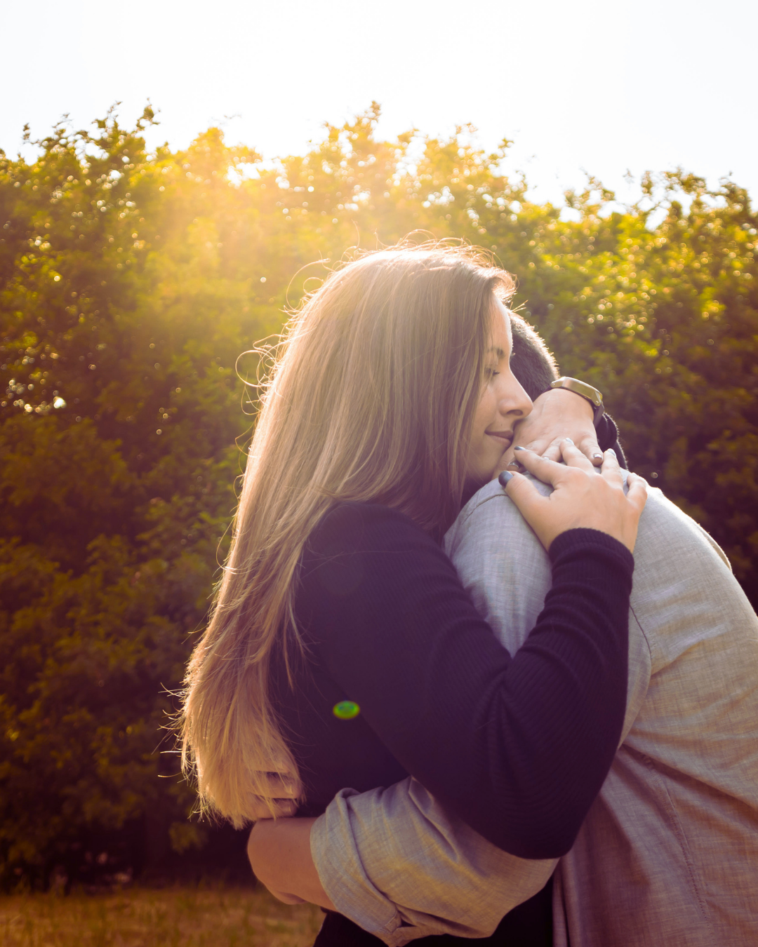 A woman with long hair hugging a person outdoors during sunset, with trees in the background.