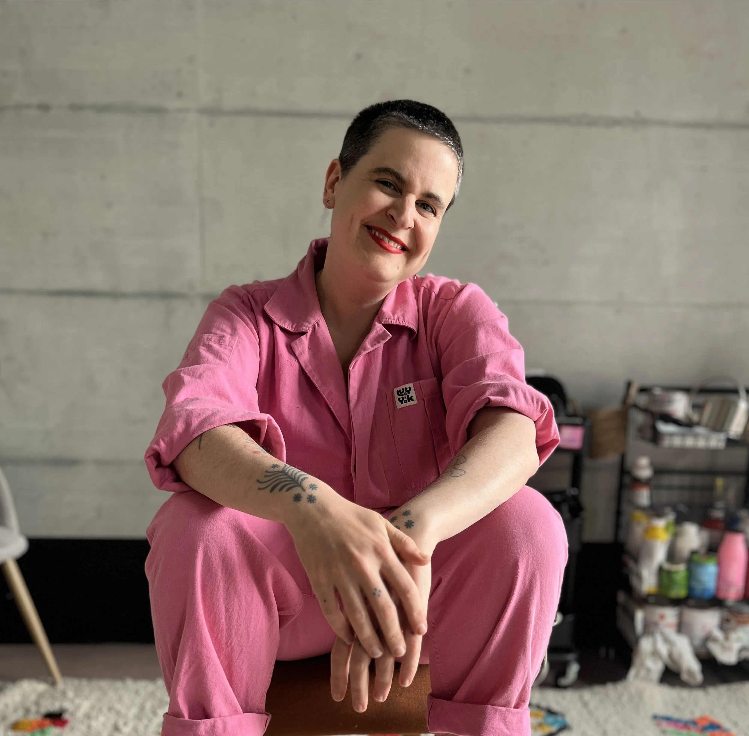 Eine Person mit kurzer schwarzer Frisur lächelt und trägt eine pinkfarbene Jacke. Sie sitzt auf einem Tisch in einem Raum mit grauer Wand. Im Hintergrund sind Küchenutensilien und Pflanzen sichtbar.