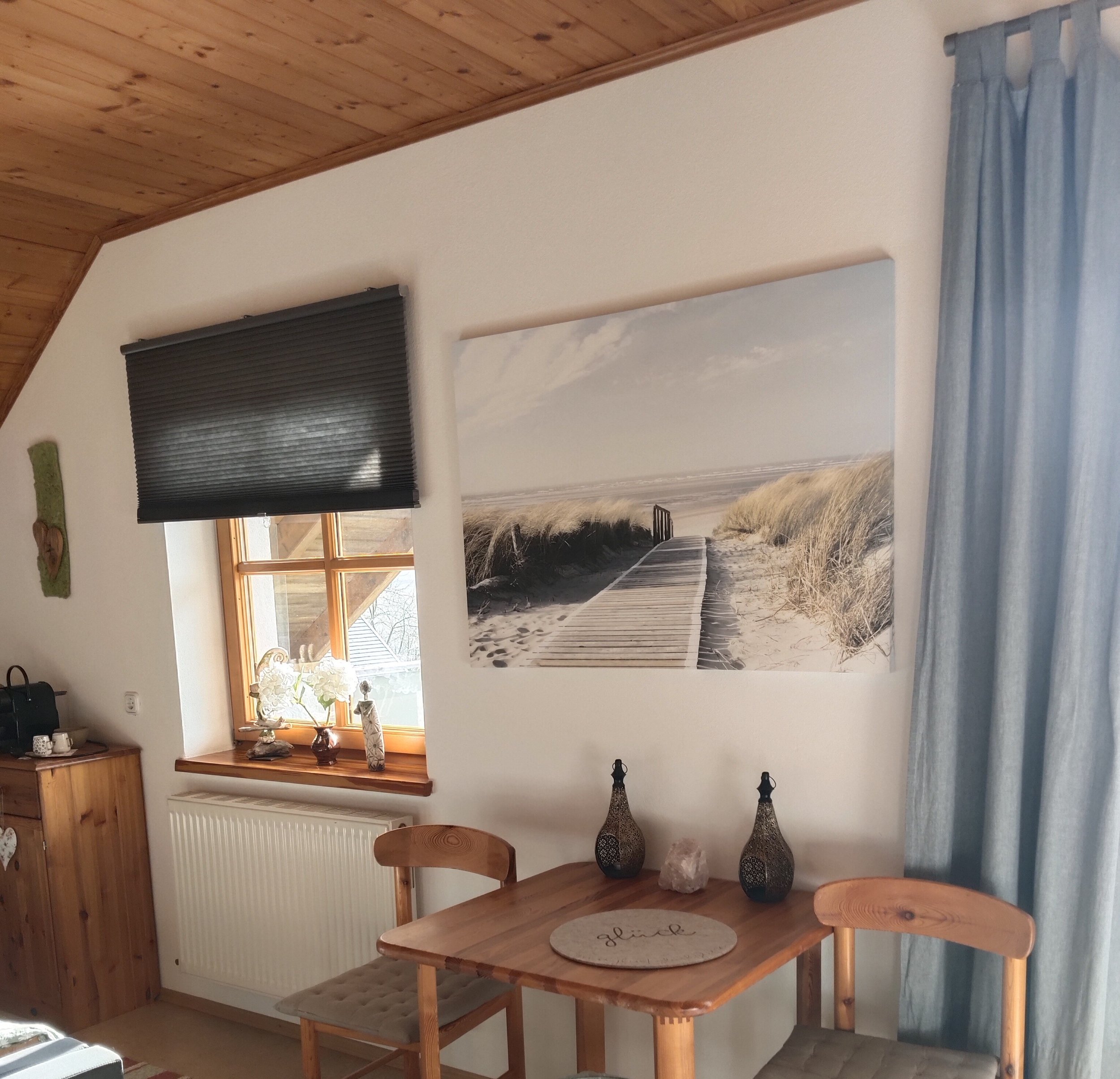Innenraum mit Holzdecke, einer kleinen Fensterbank mit Dekoration, einem Wandbild vom Strand, einem kleinen Tisch mit zwei Stühlen, Keramikvasen und einem Kristall.